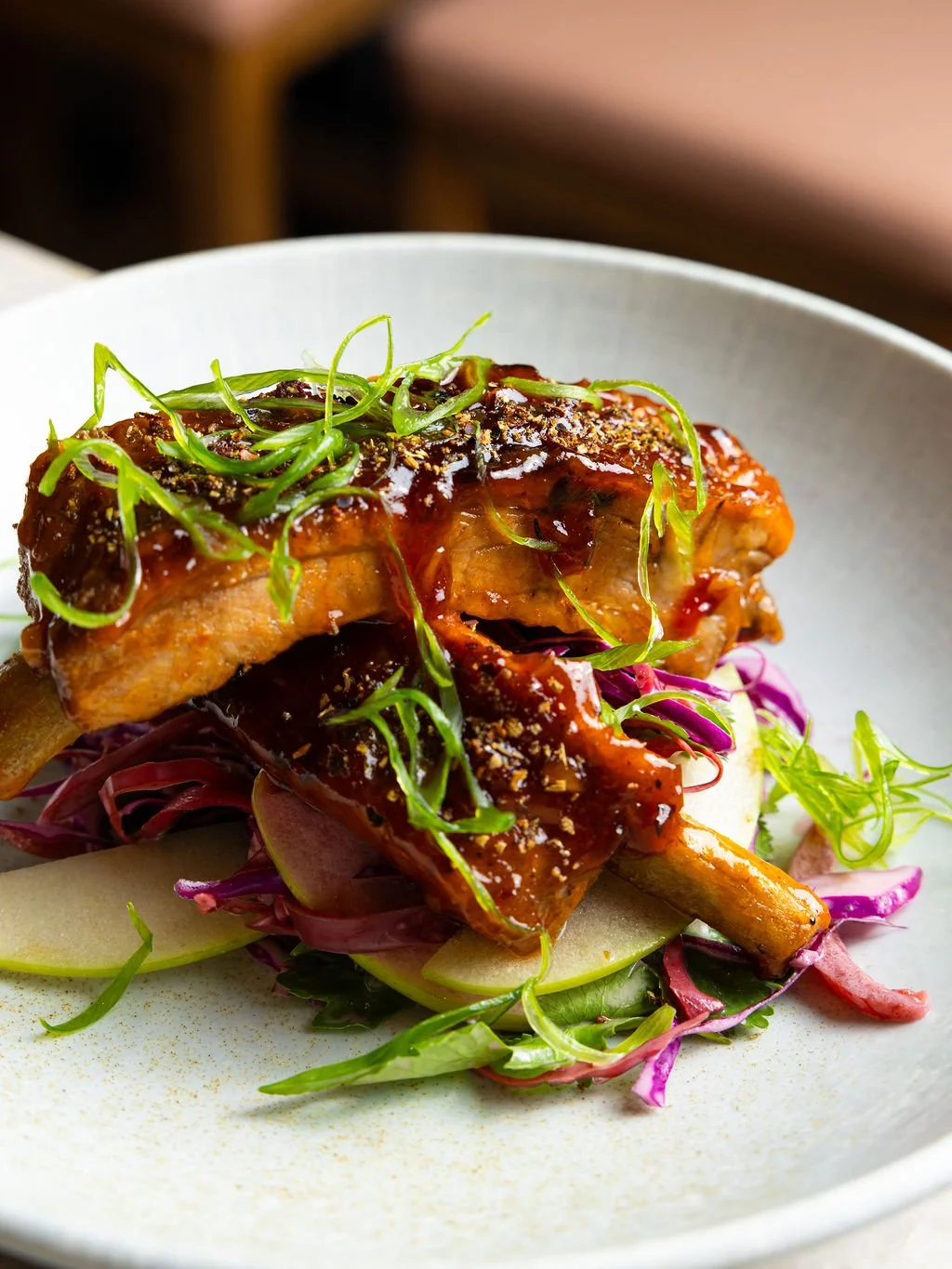 Grilled pork chop garnished with green herbs, served over a bed of mixed salad greens and sliced apples on a white plate.