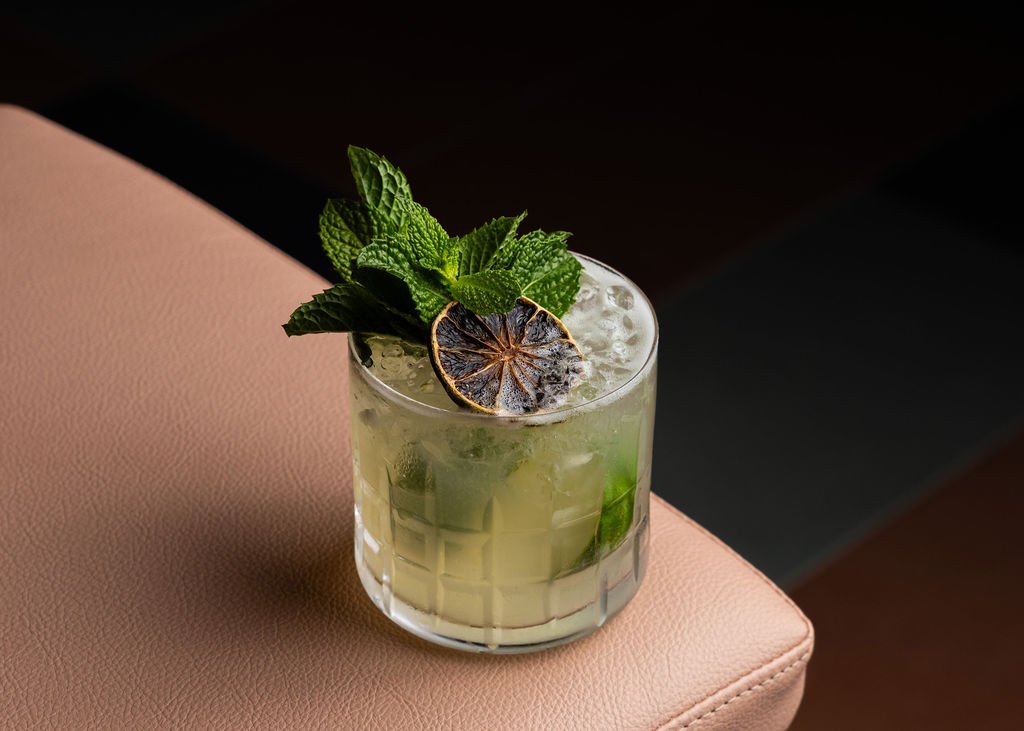 A glass of mojito cocktail with mint leaves and a slice of lime, placed on a pink upholstered surface.