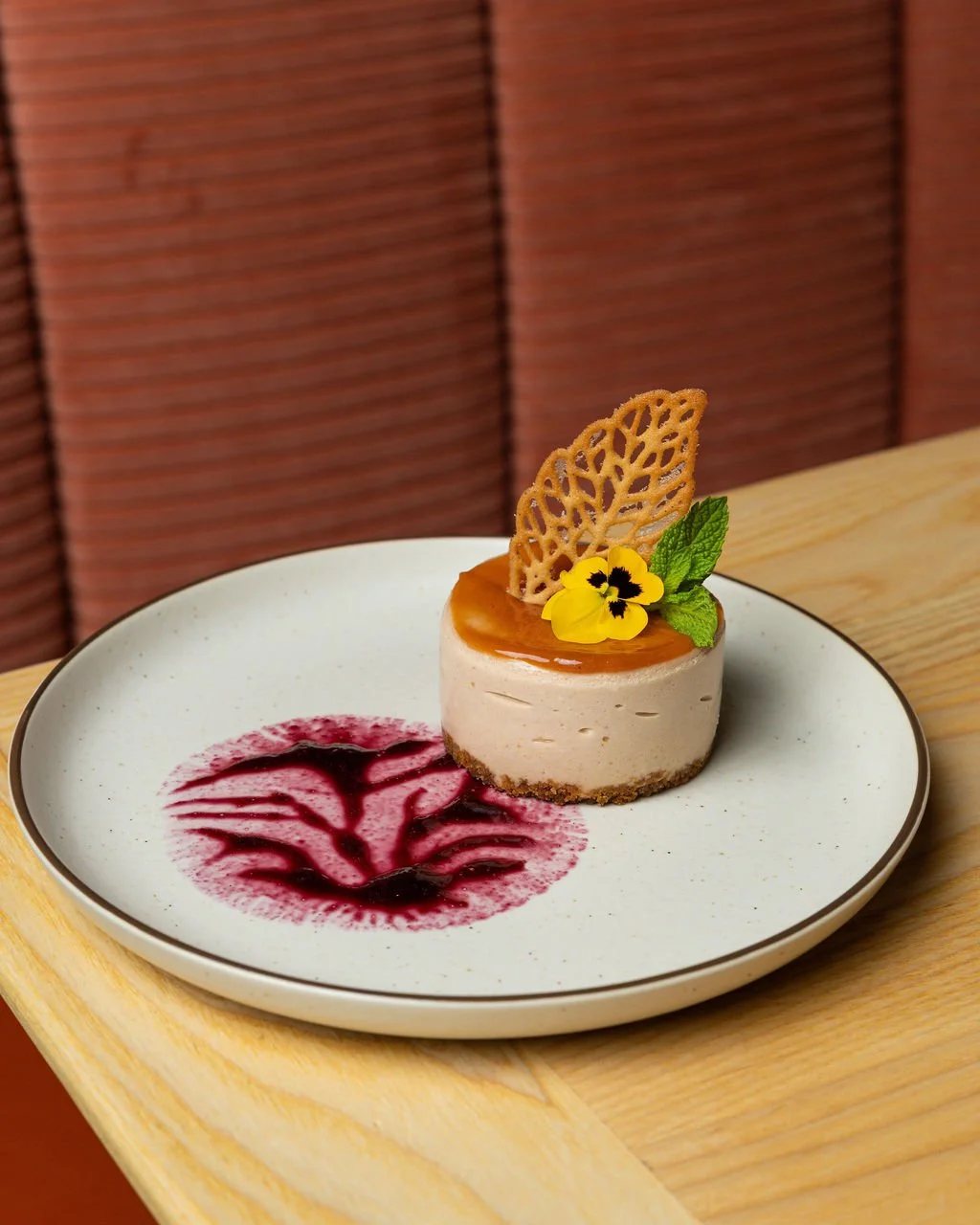 A dessert on a white plate with a pinkish-red fruit sauce design. The dessert is a small, round cheesecake topped with caramel, a decorative caramel leaf, a yellow flower, and a green mint leaf. The background features a wooden table and a cushioned 