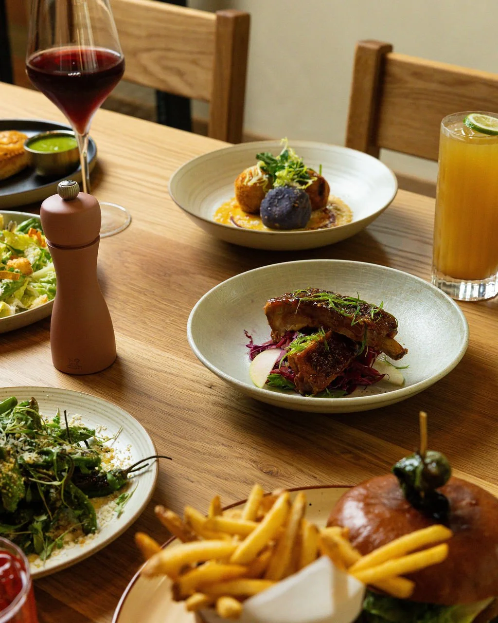 A dining table with various dishes including a salad, a plate of ribs, a bread roll with olives, and two drinks, one red wine and one citrus beverage with lime.