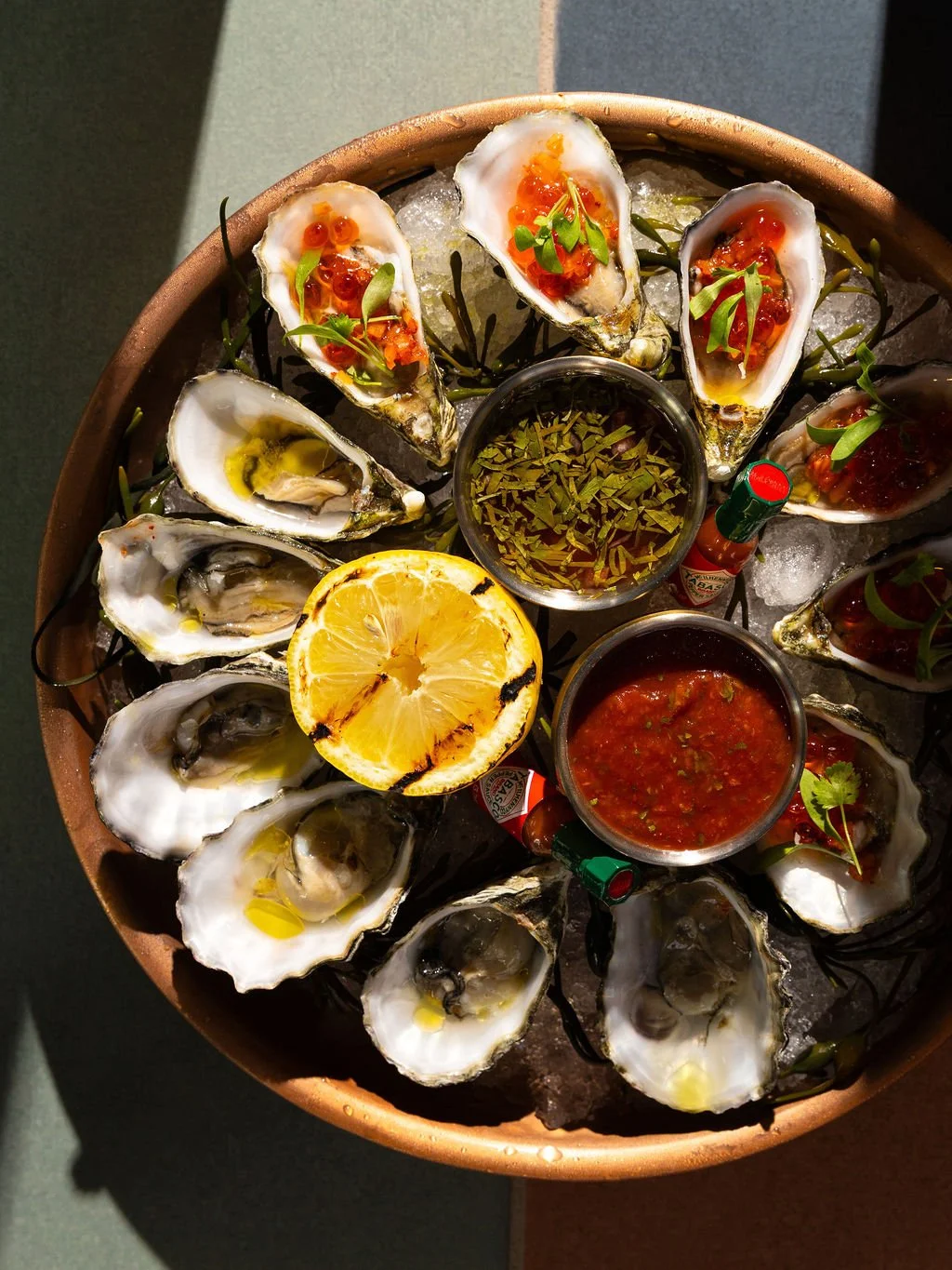 A round platter of raw oysters on ice, garnished with herbs, lemon slices, and accompanying sauces including cocktail sauce and hot sauce.