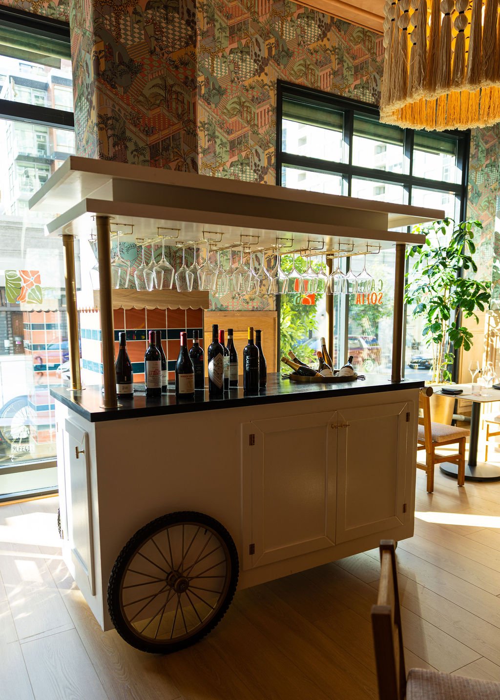 A mobile wine cart with bottles of wine and upside-down wine glasses hanging above, set in a brightly lit restaurant with patterned wallpaper and large windows letting in natural light.