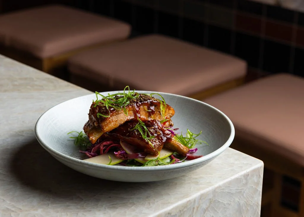 A white bowl of glazed ribs with green garnish, placed on a light-colored marble or stone table.
