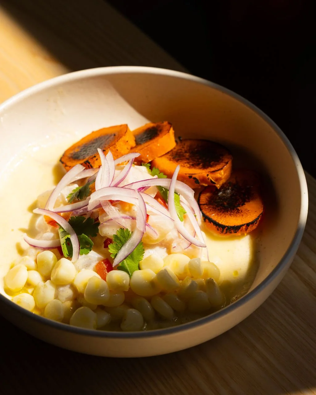 A bowl of grilled sweet potato slices with charred marks, topped with sliced red onions, fresh cilantro, and a mixture of corn kernels and diced vegetables in a creamy sauce.