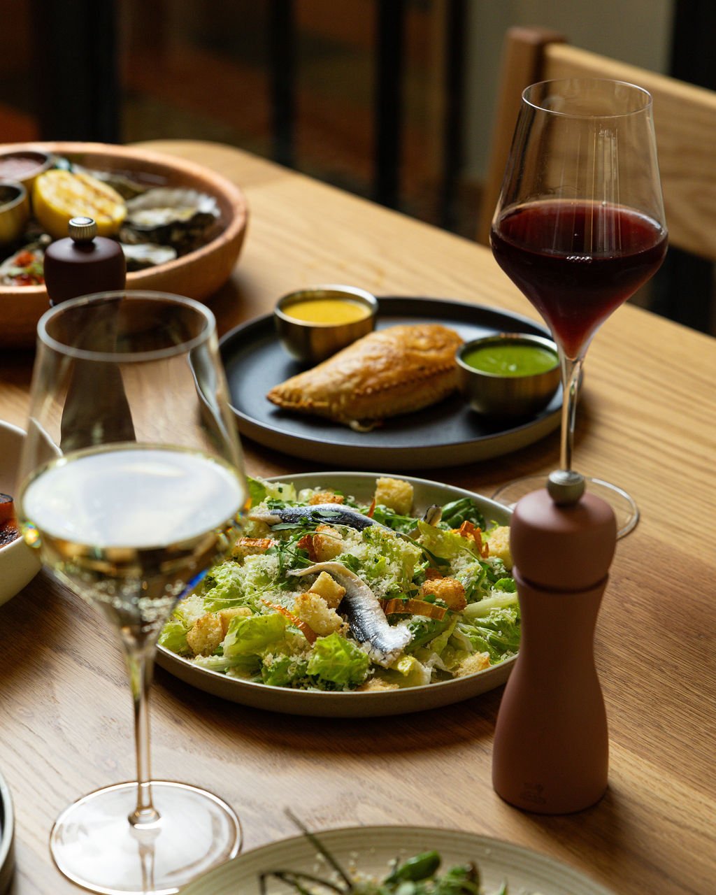 A wooden table set with various dishes, including a glass of white wine, a glass of red wine, a salad with anchovies, a plate with a piece of fish and sauces, and a bowl of oysters in the background.
