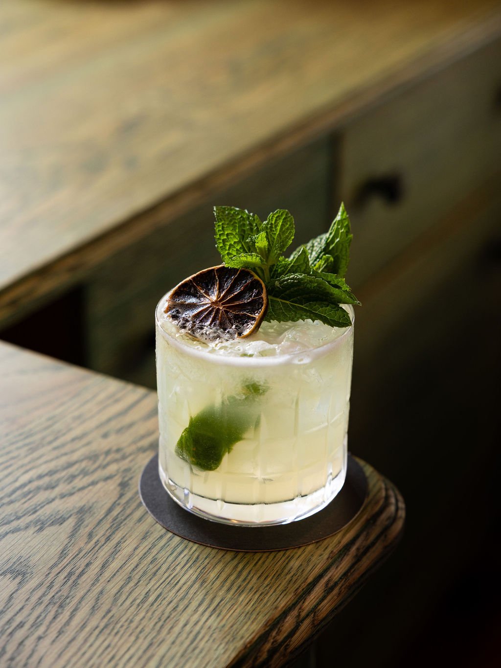 A refreshing cocktail with mint leaves and a dried lemon slice garnished on top, served in a clear glass with ice and lime wedges inside, placed on a black coaster on a wooden surface.