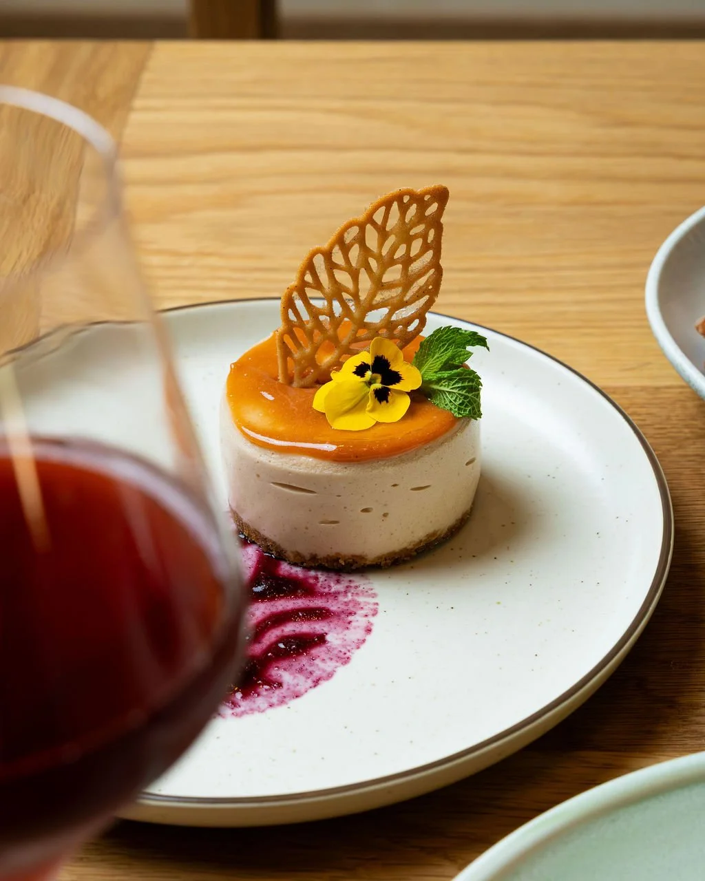 A plated dessert featuring a white mousse cake topped with a caramel glaze, garnished with a decorative lattice, a yellow edible flower, and a green mint leaf, with a red sauce smear on the plate.