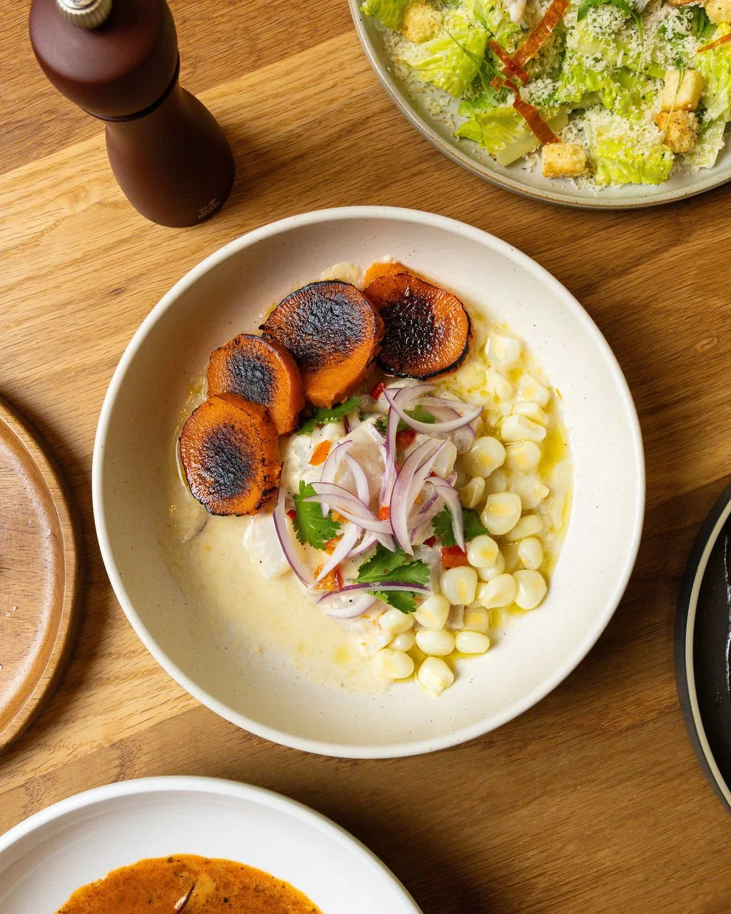 A bowl of roasted sweet potatoes, topped with onions, fresh cilantro, and accompanied by corn, served on a wooden table.