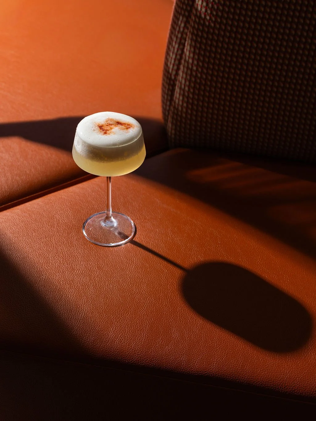 A cocktail with foam and a sprinkle of cinnamon in a coupe glass on a textured orange surface, with a shadow cast on the surface.
