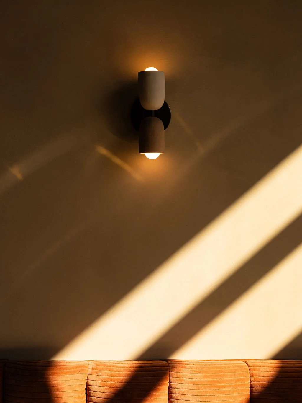 A ceiling light fixture with two light bulbs casting shadows on the ceiling, above an orange striped sofa with sunlight streaming through blinds.