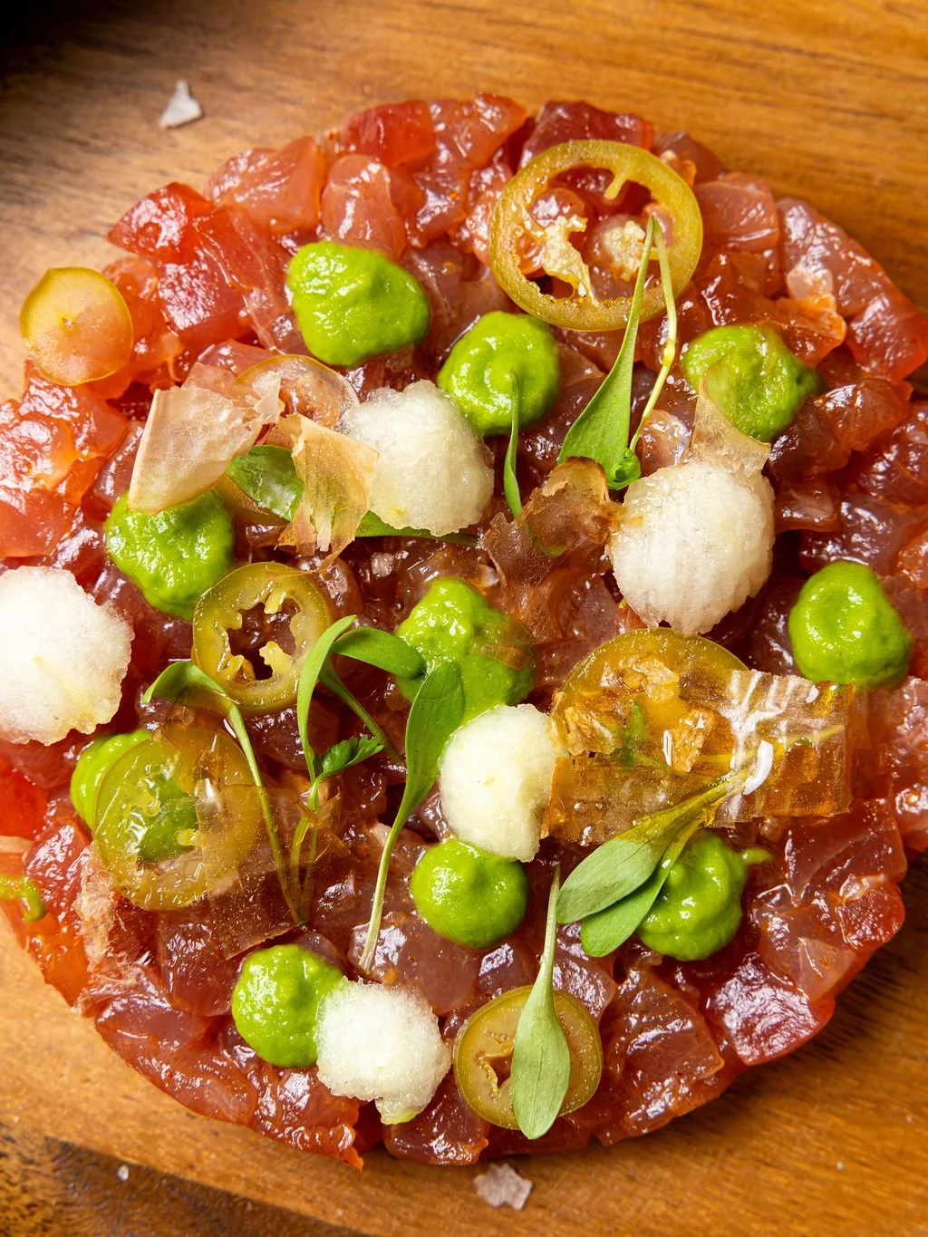 Close-up of a dish with diced raw fish, green peas, sliced jalapenos, foam, and microgreens on a wooden surface.