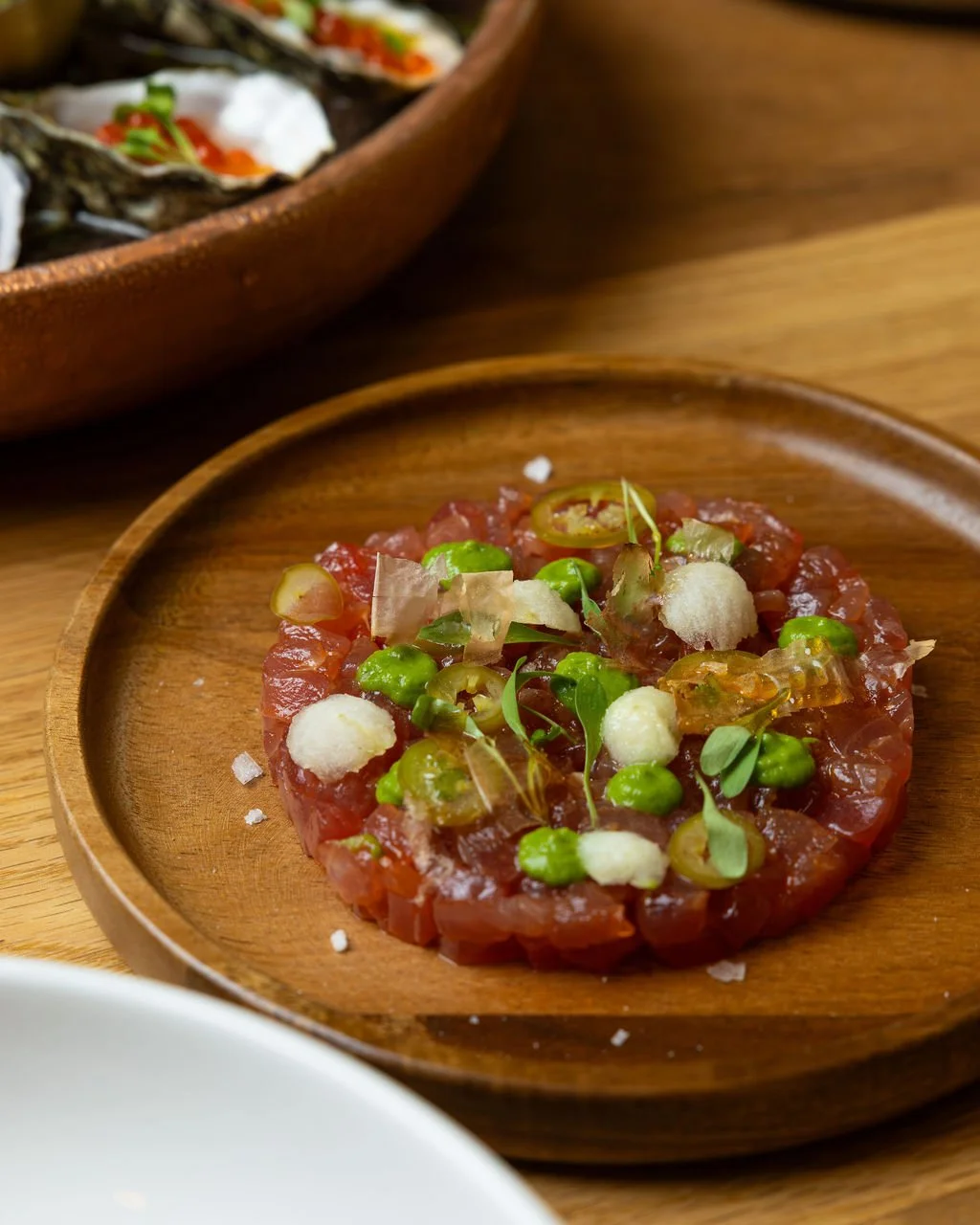 Close-up of a carne tartare on a wooden plate, garnished with microgreens, sliced jalapeños, and small white and green dollops, with a larger bowl of similar dish in the background.