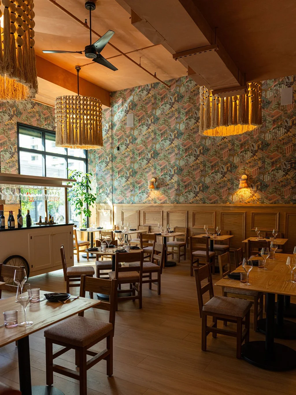 Interior of a restaurant with wooden tables and chairs, colorful geometric patterned wallpaper, large hanging woven light fixtures, and a ceiling fan. Features a window with natural light and some greenery outside.