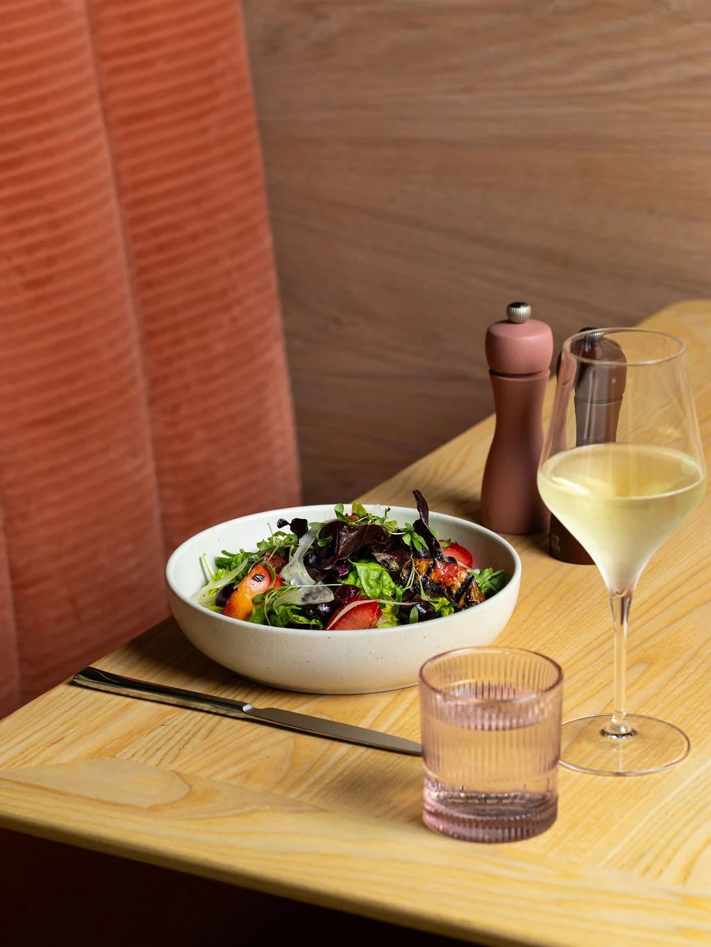 A dining table with a bowl of salad, a glass of white wine, a pink glass, a pink salt grinder, and a knife.