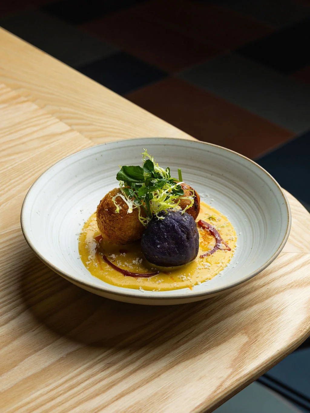 A bowl of gourmet food featuring a yellow sauce, two fried balls, one purple croquette, garnished with fresh green microgreens, on a wooden table.