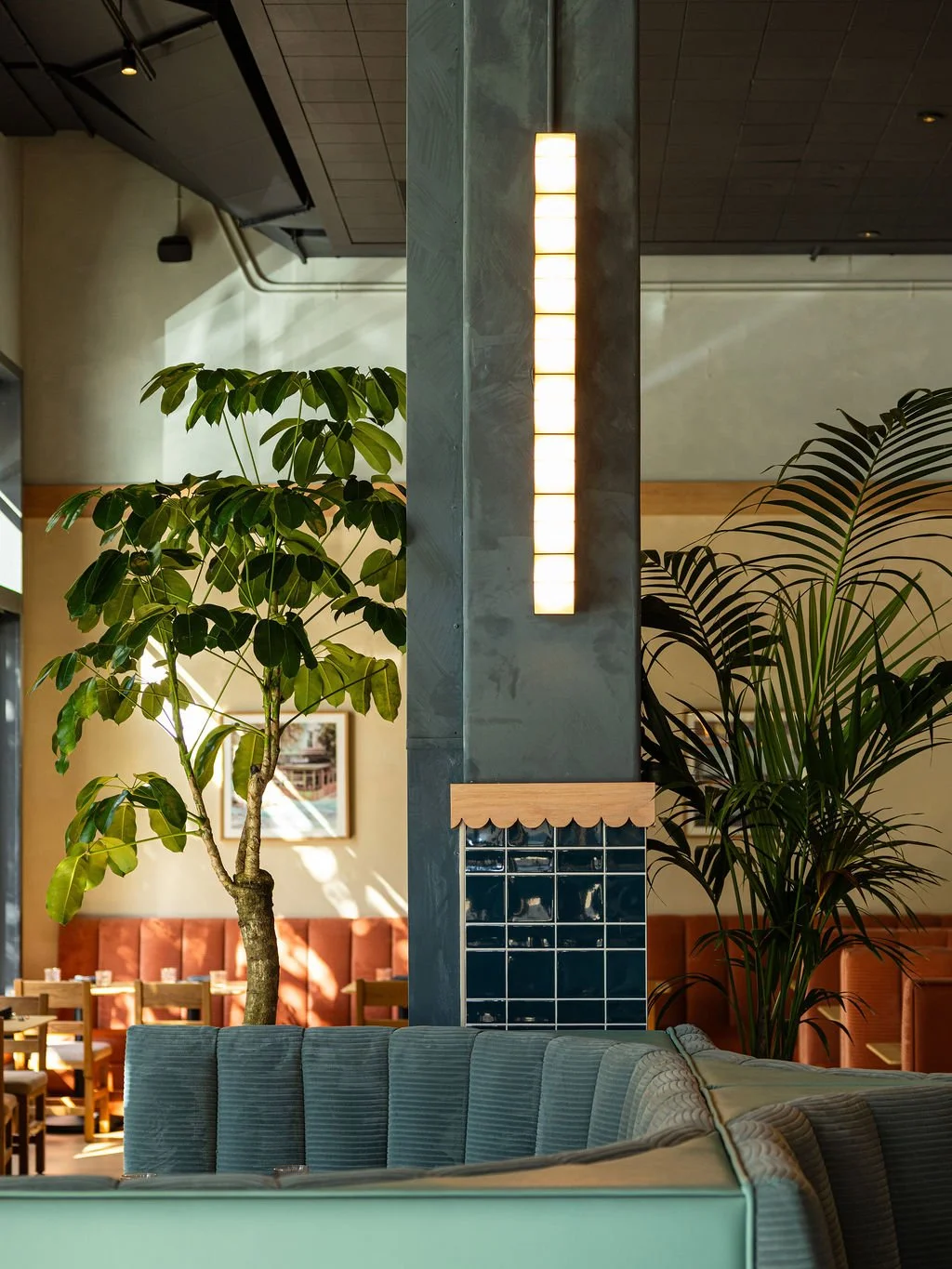 Indoor restaurant with large potted plants, a seating area with a light green sofa, and a modern wall-mounted light fixture.