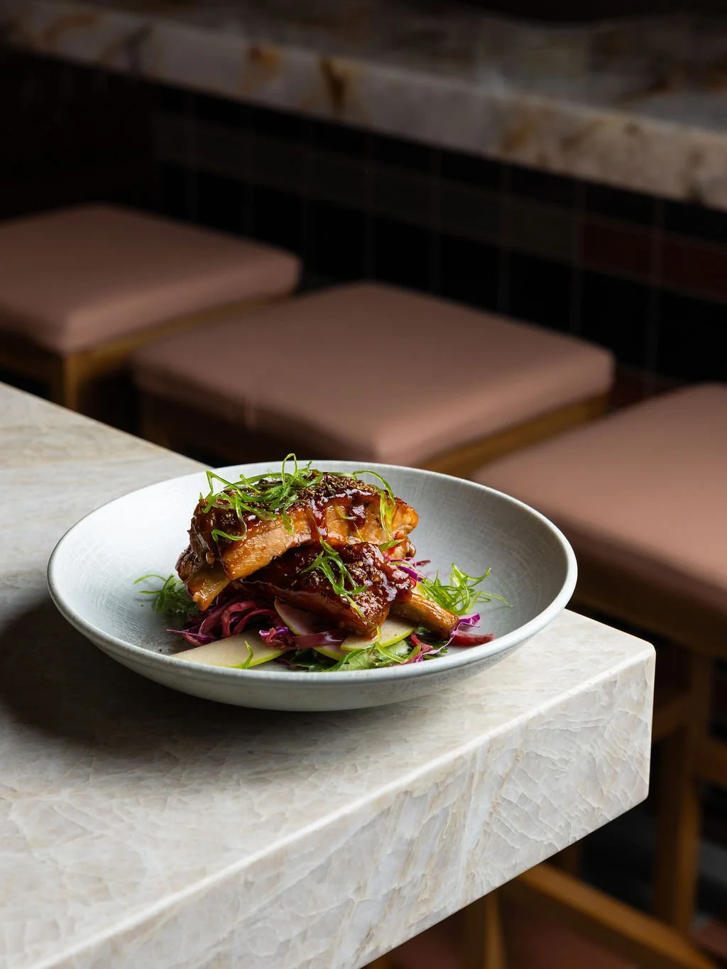 A white plate of glazed meat on a bed of purple and green vegetables sits on a marble countertop in a dimly lit restaurant setting.