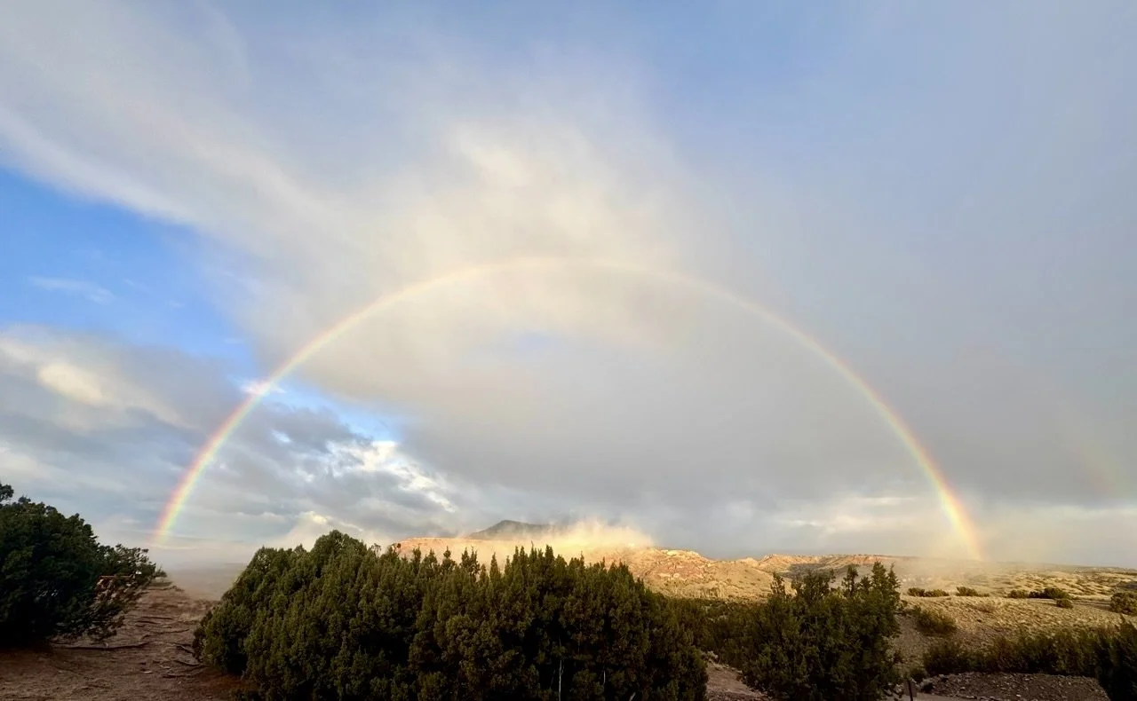 A double rainbow arcs over a desert landscape with shrubs and distant hills, partly covered by clouds and blue sky. soul path integration transformational coaching hypnotherapy higher self life coach hypnotherapy human design