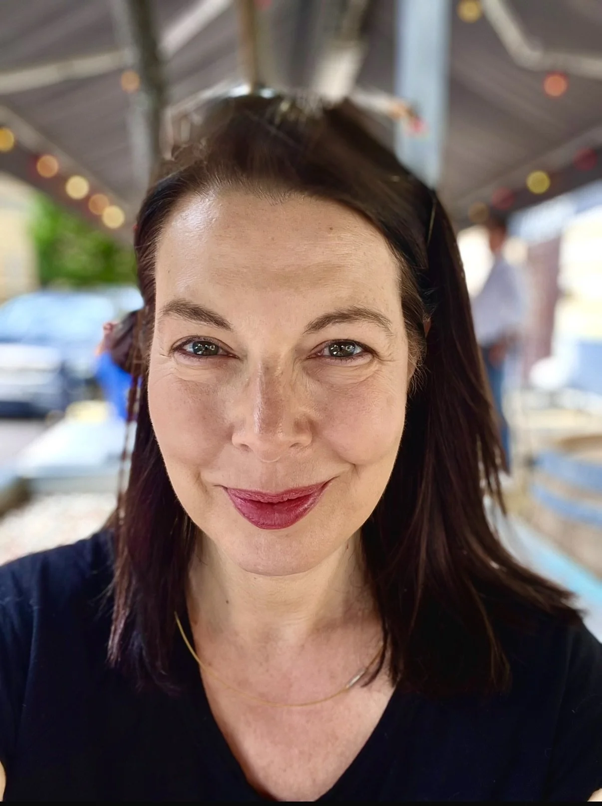 A woman with dark brown hair, wearing red lipstick and a black top, taking a close-up selfie under a canopy with people and cars in the blurred background.
