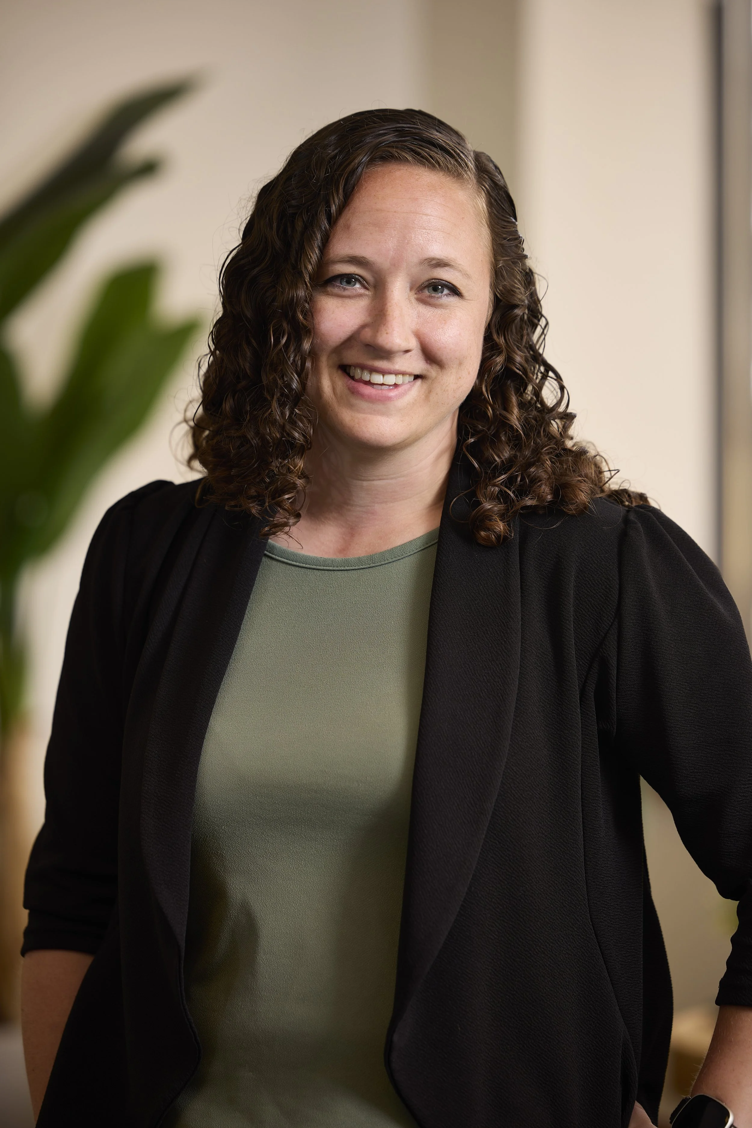 A woman with shoulder-length curly brown hair, wearing a green top and black blazer, smiling and standing indoors.