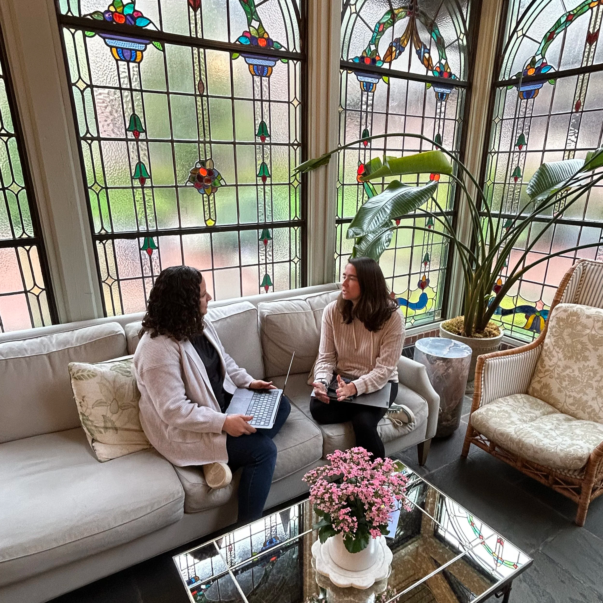 Two women having a conversation in a cozy, well-lit room with large stained glass windows, a white sofa, a glass coffee table with pink flowers, a tall potted plant, and an armchair.