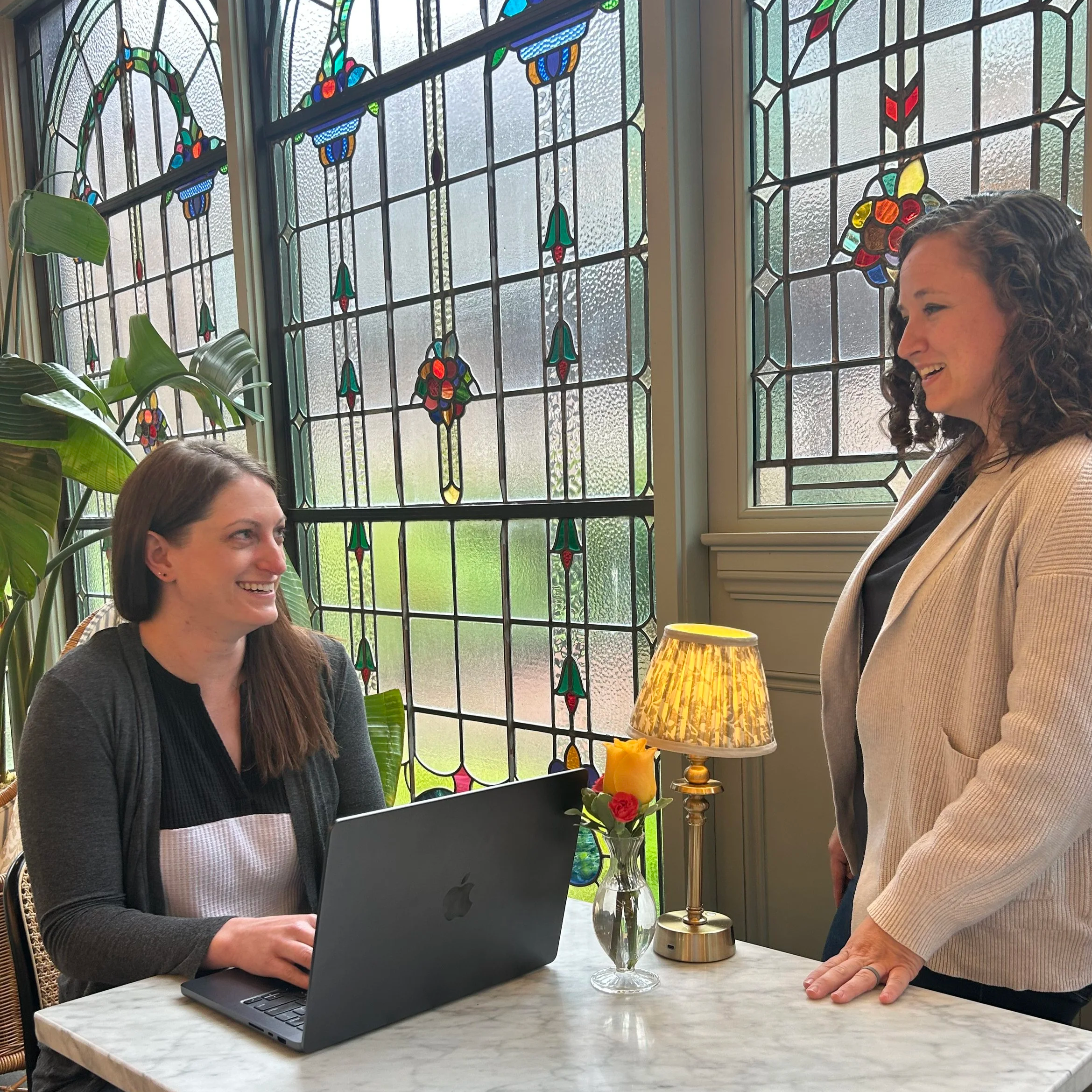 Two women are smiling and talking in a bright room with stained glass windows. One woman is sitting at a marble table with a laptop, and the other woman is standing beside the table. There is a small lamp and a vase with a yellow and red flower on the table.
