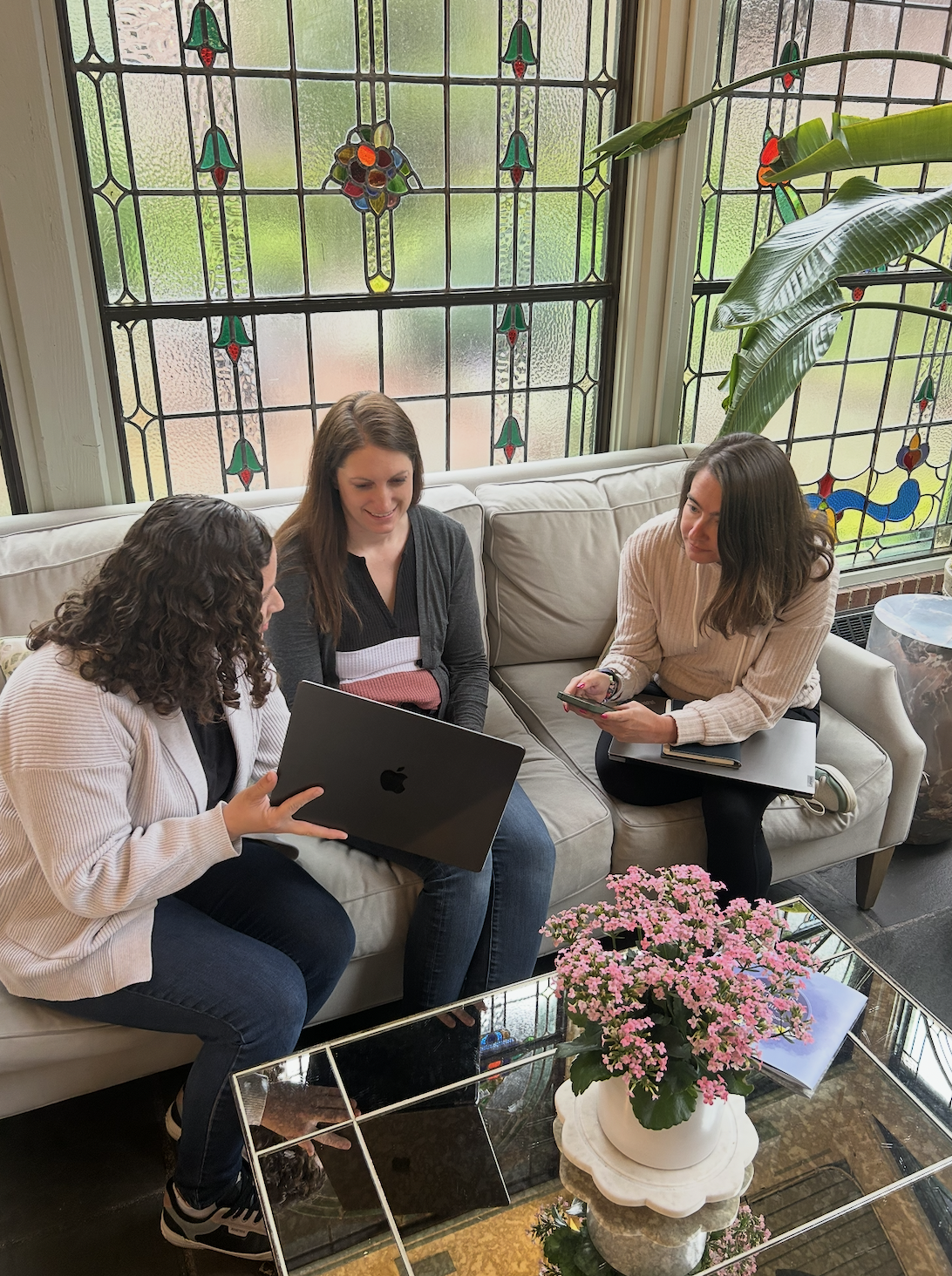 Three women sitting on a beige sofa in a room with stained glass windows, discussing work while one woman shows a laptop and another holds a phone, with pink flowers on a glass coffee table in front of them.