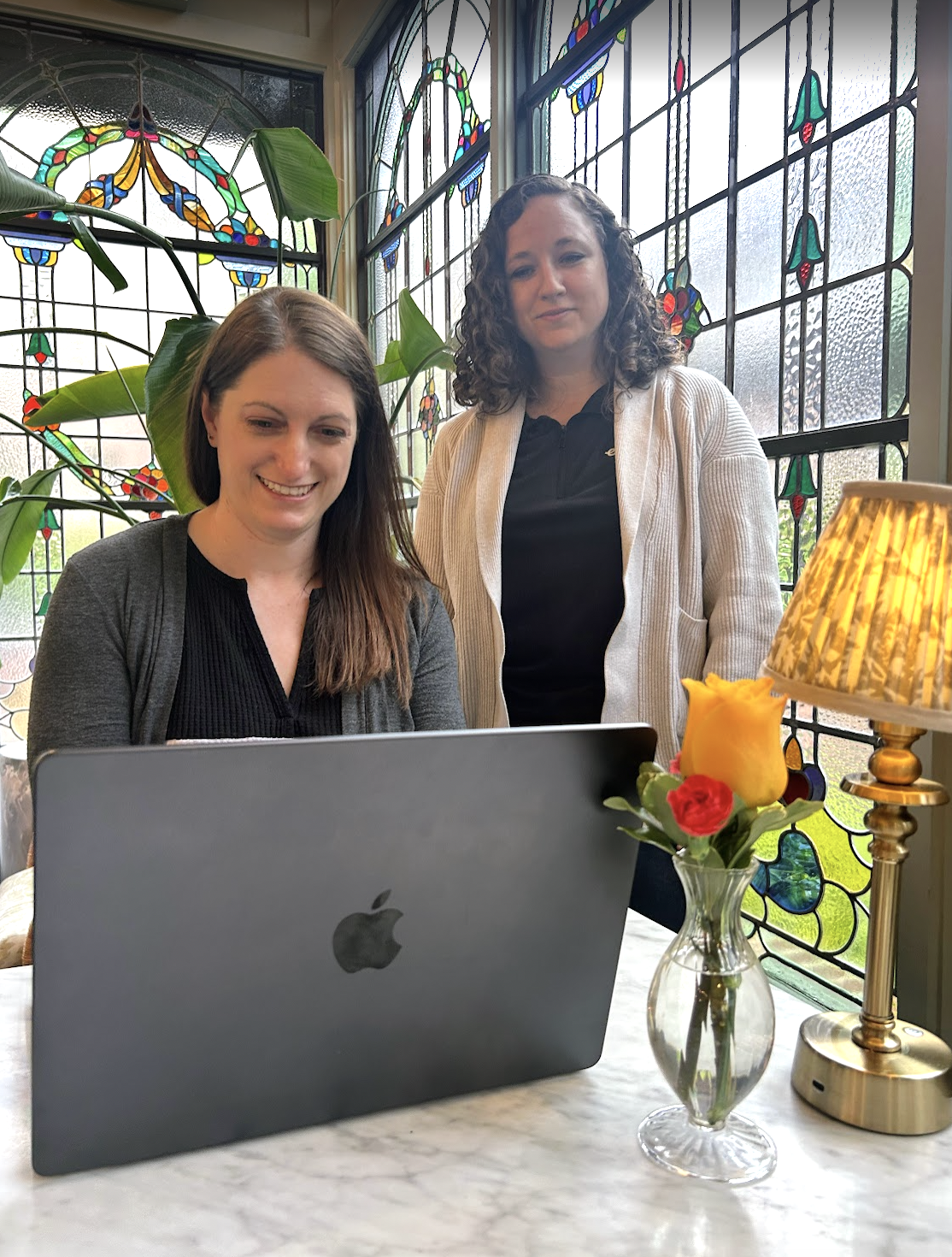 Two women looking at a laptop on a marble table, surrounded by stained glass windows, plants, a table lamp, and a vase with yellow and red roses.