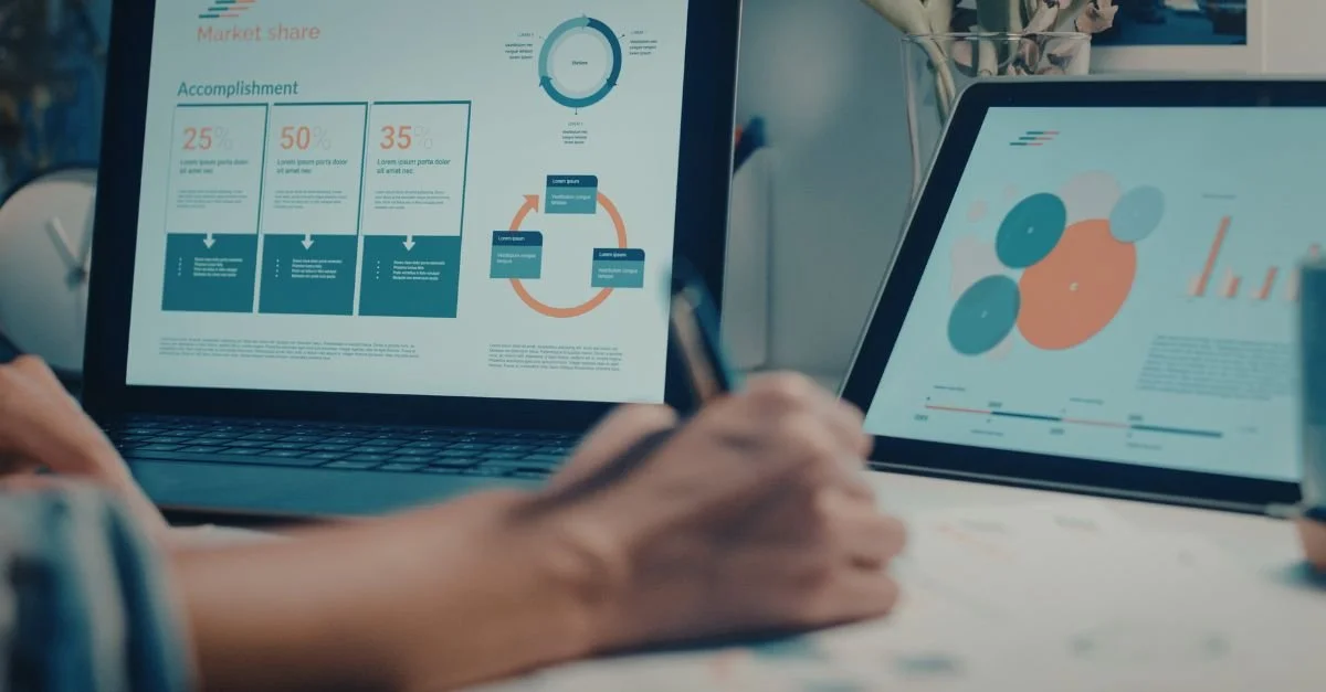 Two screens display business charts and graphs, with a person taking notes at a table.