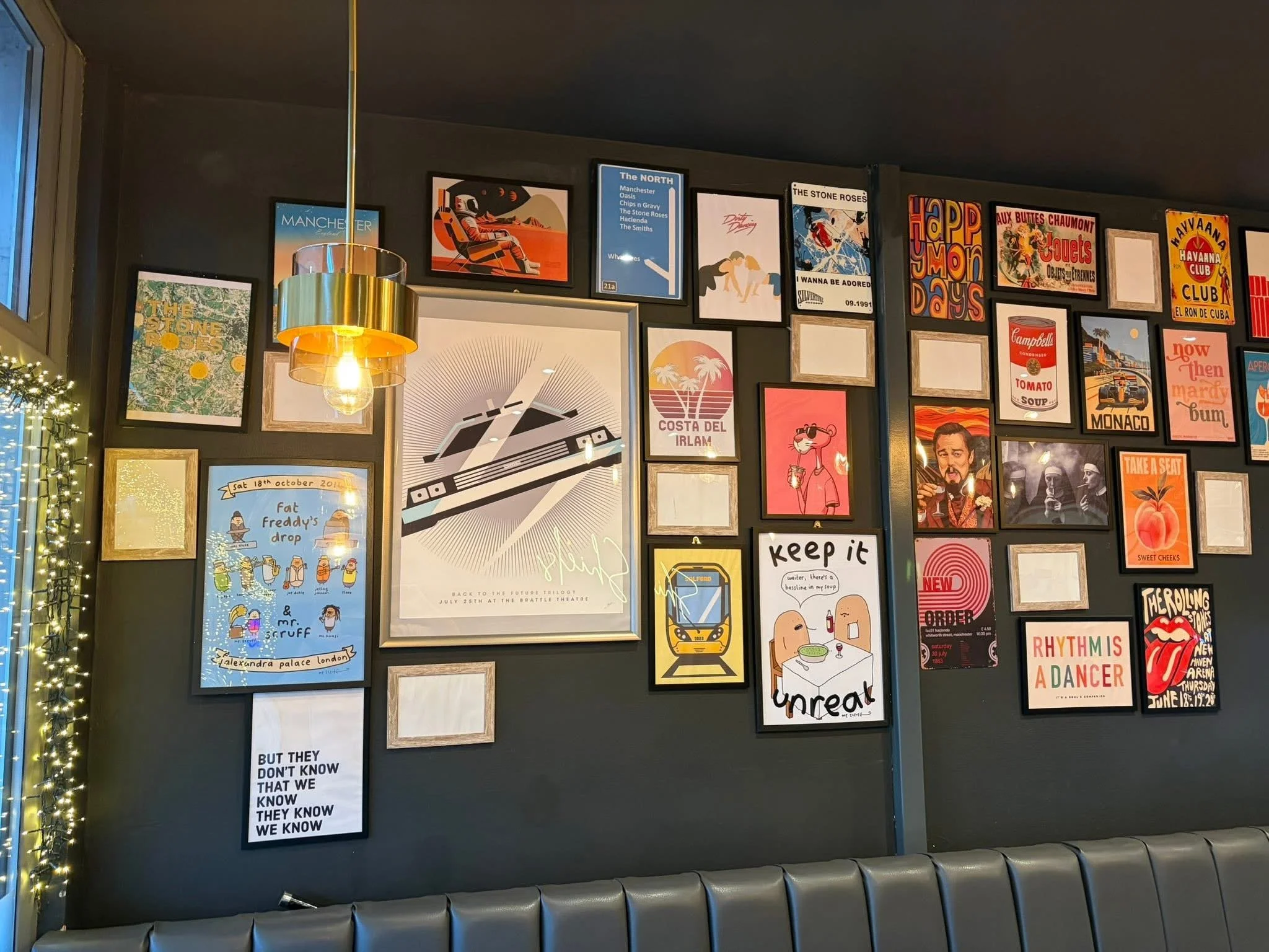 A gallery wall with various framed posters and artwork in a cafe or restaurant, decorated with string lights on the left side, and a gray upholstered seating along the bottom.