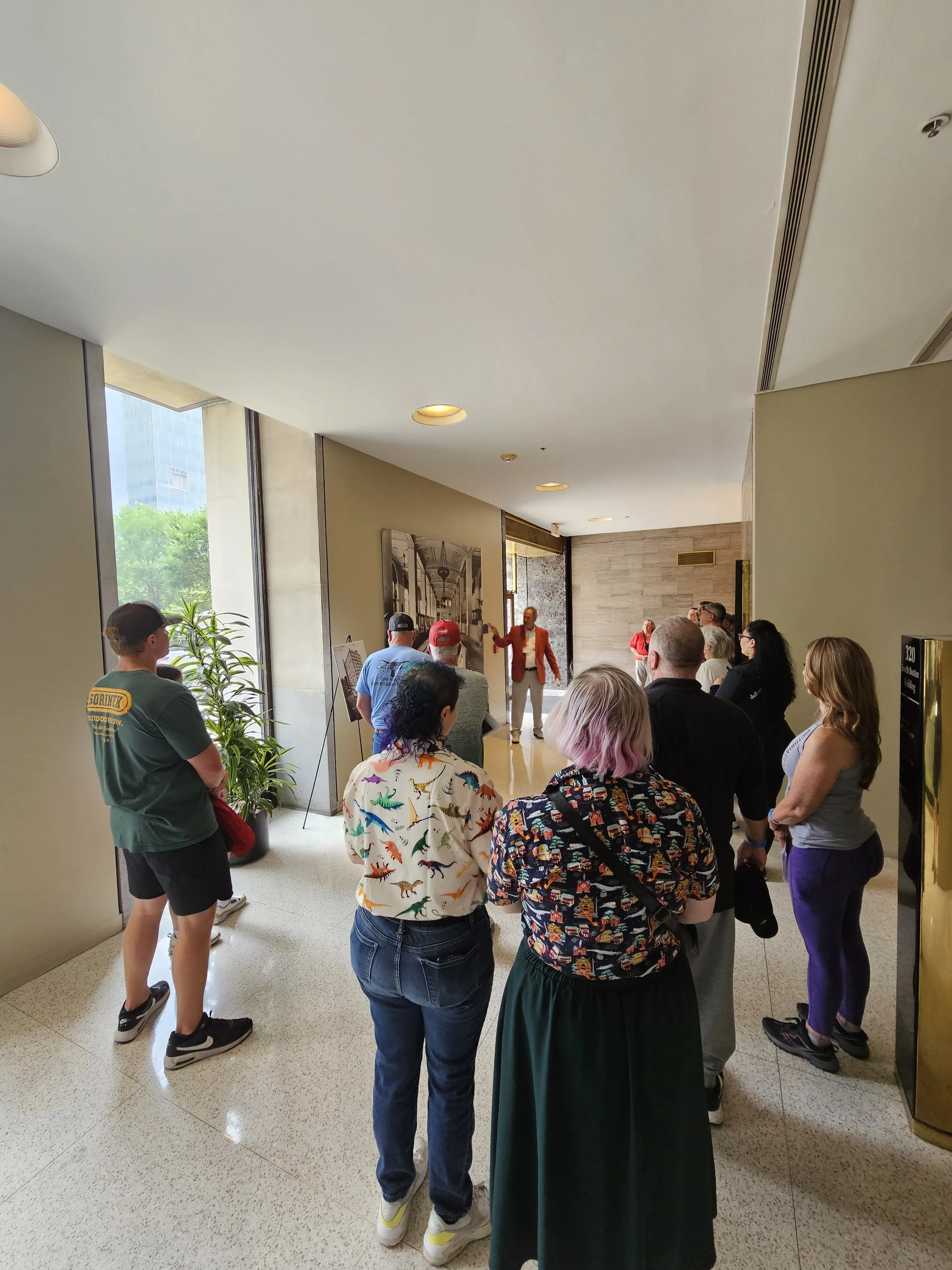 People standing in a lobby or hallway, listening to a man in an orange blazer speaking to the group, with large windows and framed photographs on the walls.