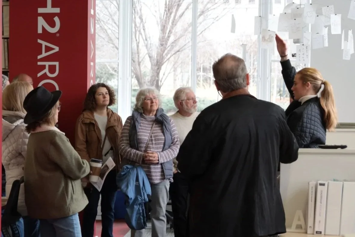 A group of people listening to a woman explaining something on a wall filled with notes inside a modern building with large windows and trees outside.