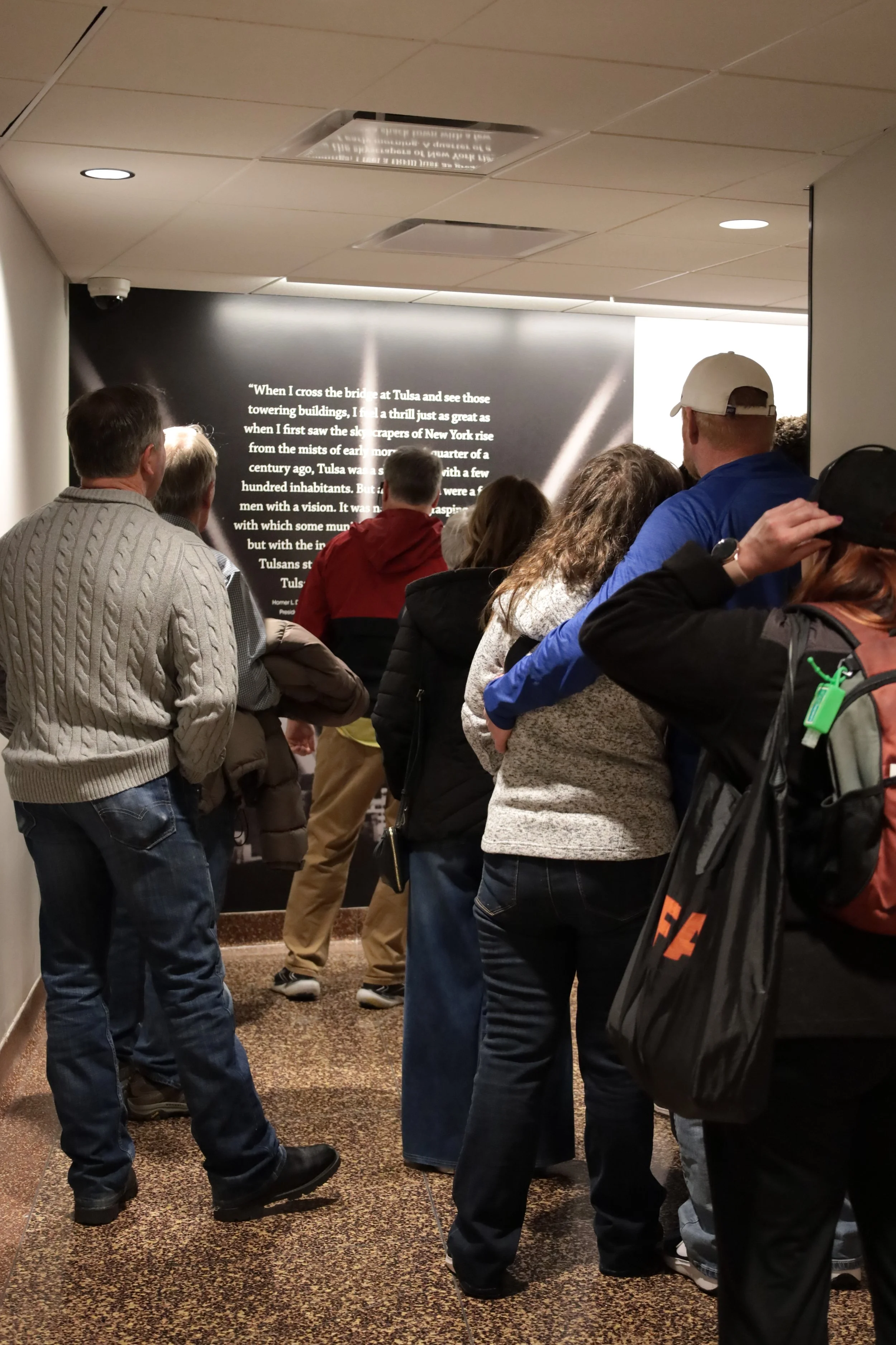 Group of people standing in line inside a museum, facing a wall with a large display of text about Tulsa and its history.