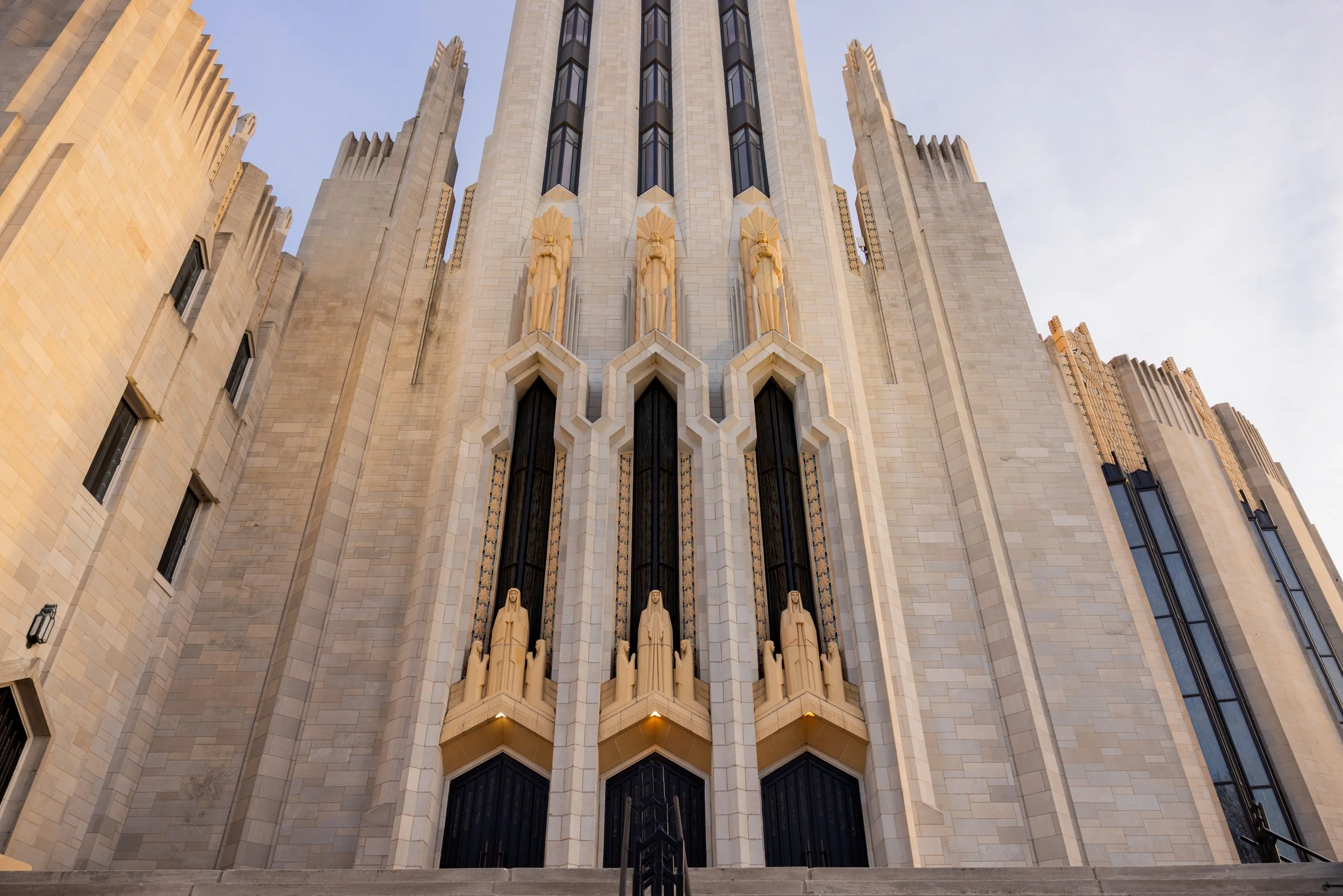 Low-angle view of a tall, ornate church building with intricate Art Deco architectural details, statues along the facade, and large stained-glass windows, under a clear sky.
