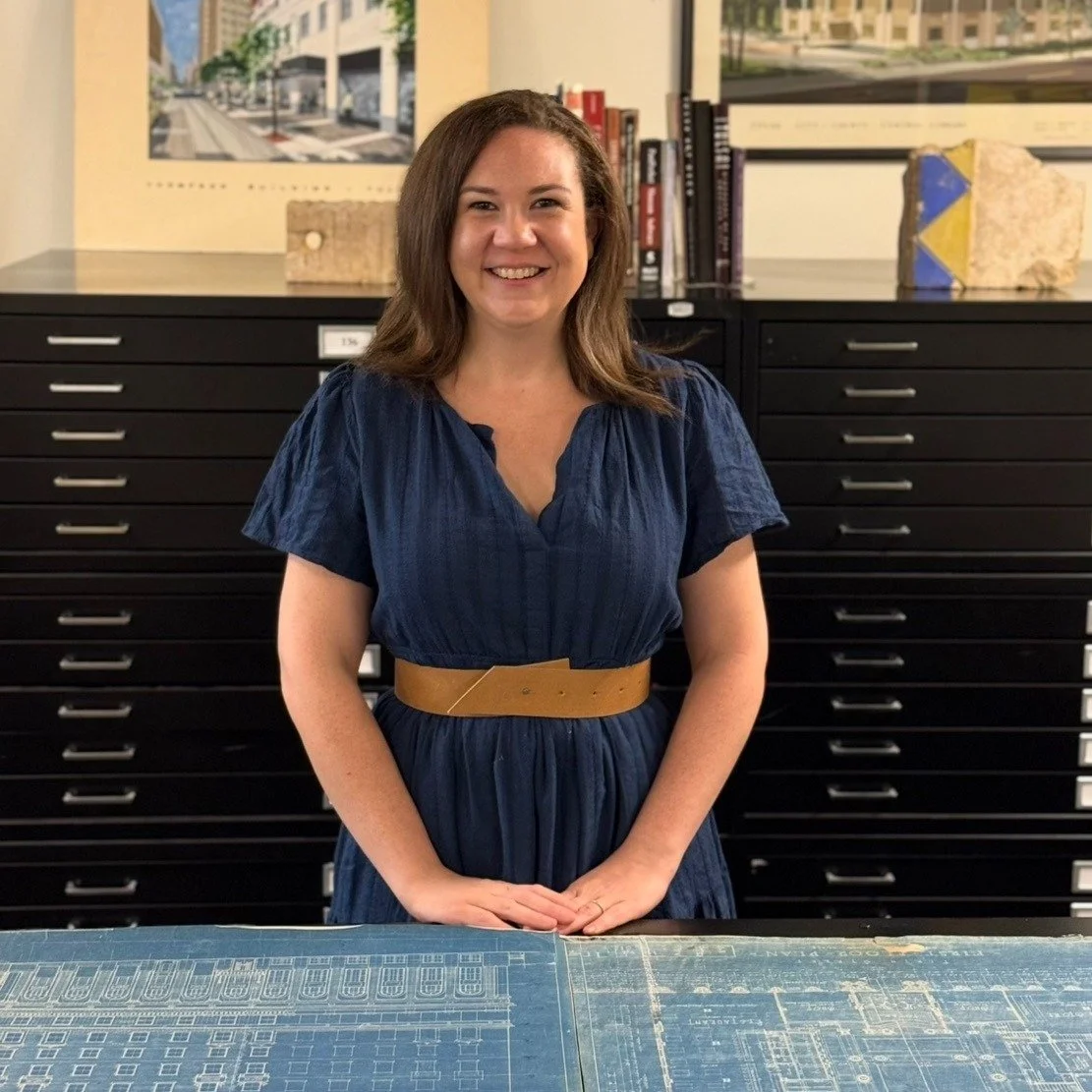 A smiling woman with shoulder-length brown hair, wearing a dark blue dress with a light brown belt, standing behind a table with blueprints in a room with black filing cabinets and artwork in the background.
