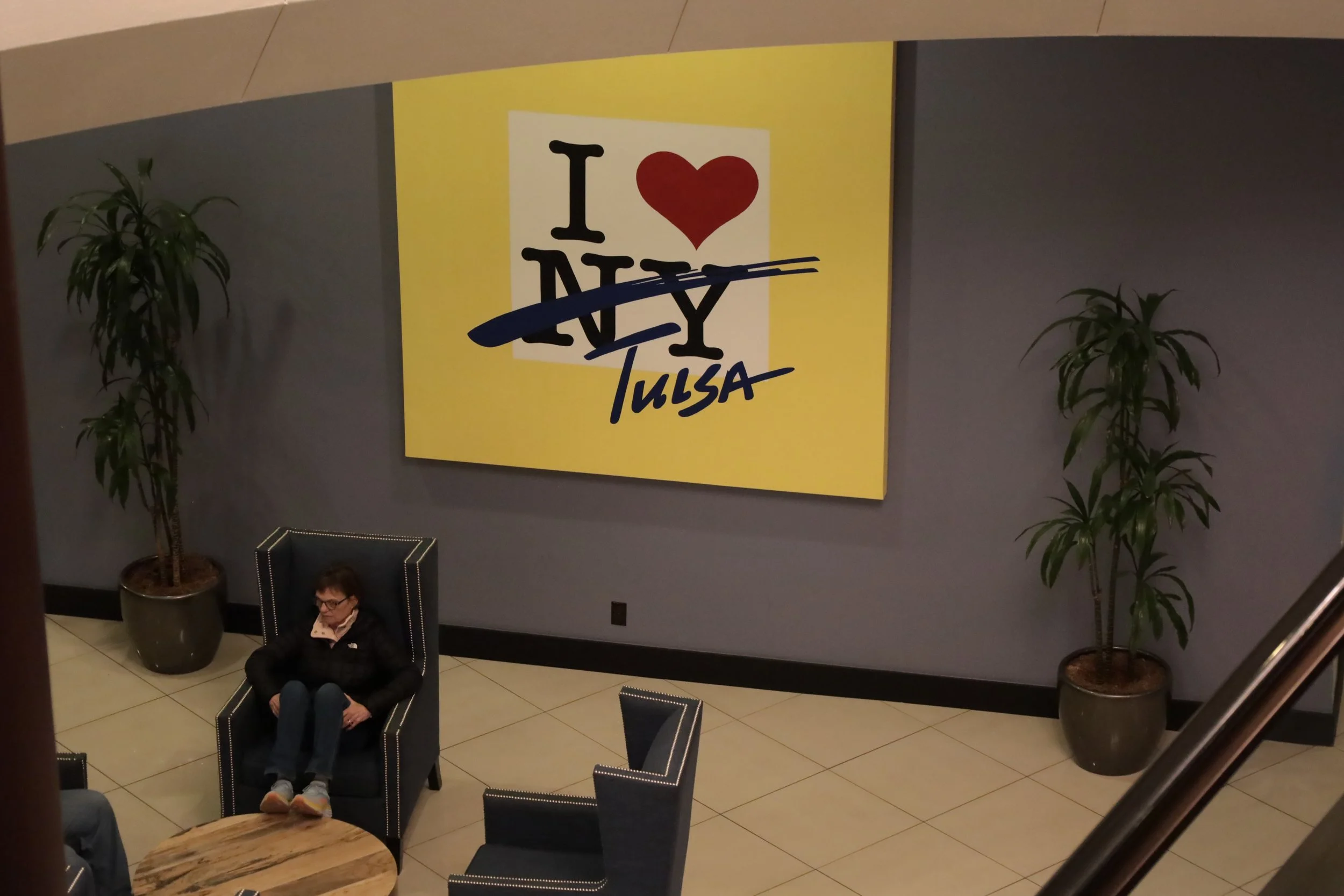 Large yellow and white sign on a gray wall that says "I ❤️ NY Tulsa" with a blue strikethrough and the word "Tulsa" handwritten in blue. Two potted plants on either side of the sign. A woman seated in a black armchair underneath the sign, with furniture including a round wooden table and another black chair visible.