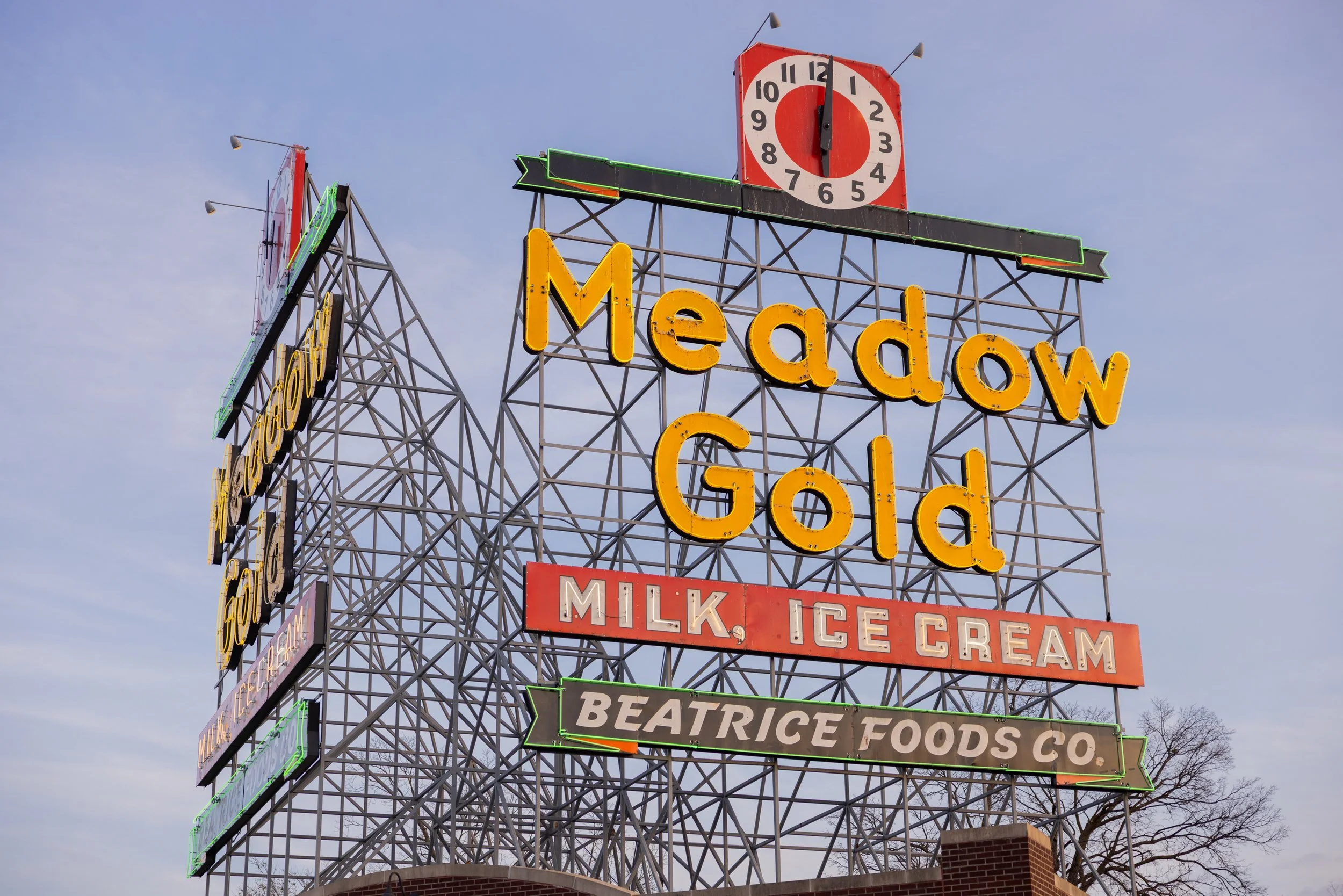 Neon sign for Meadow Gold dairy and ice cream with a clock showing 12:00, against a cloudy sky.
