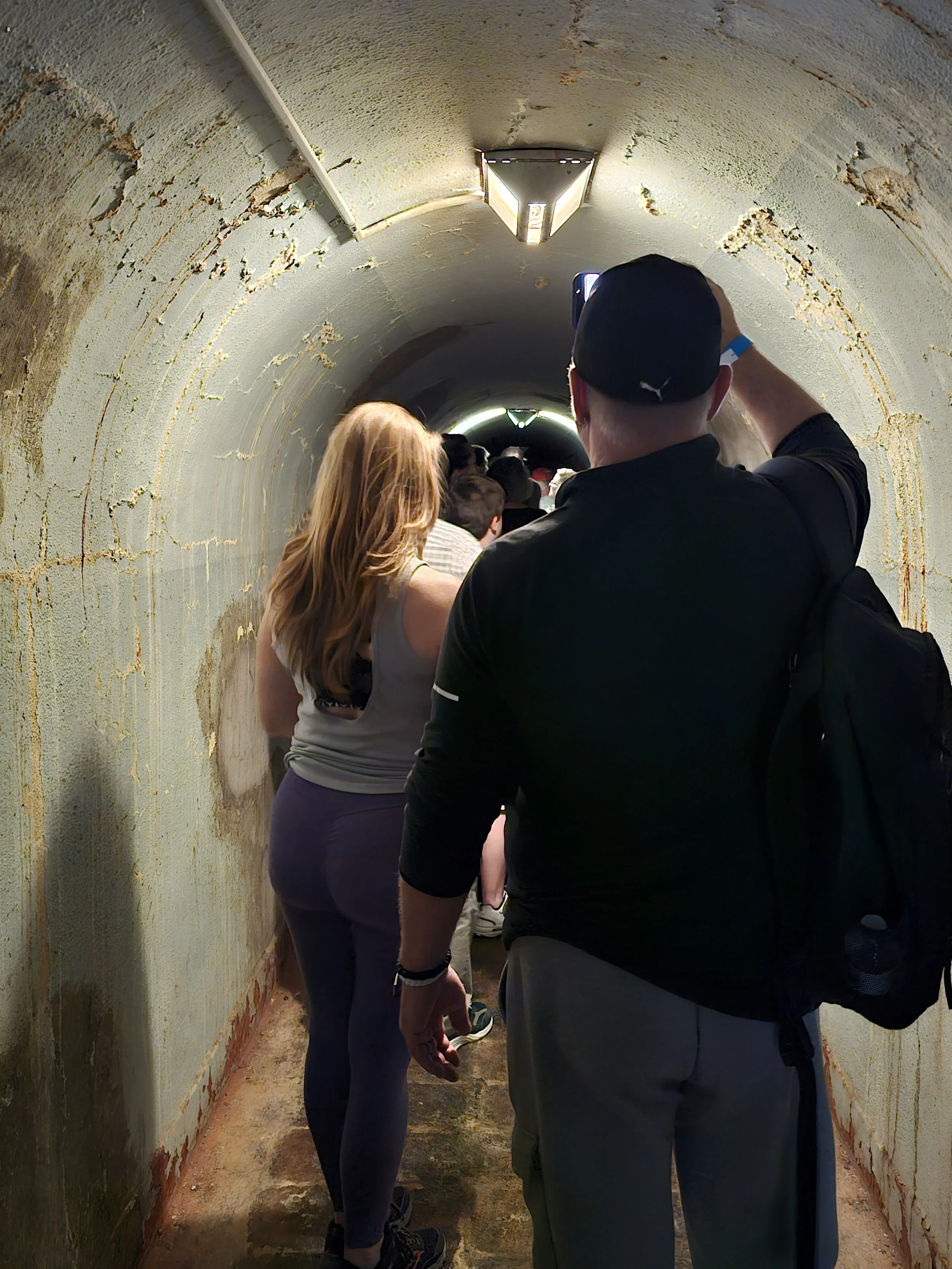 People walking in a narrow, underground tunnel with peeling, stained walls and a curved ceiling, illuminated by overhead lights. A man is taking a photo with his phone.