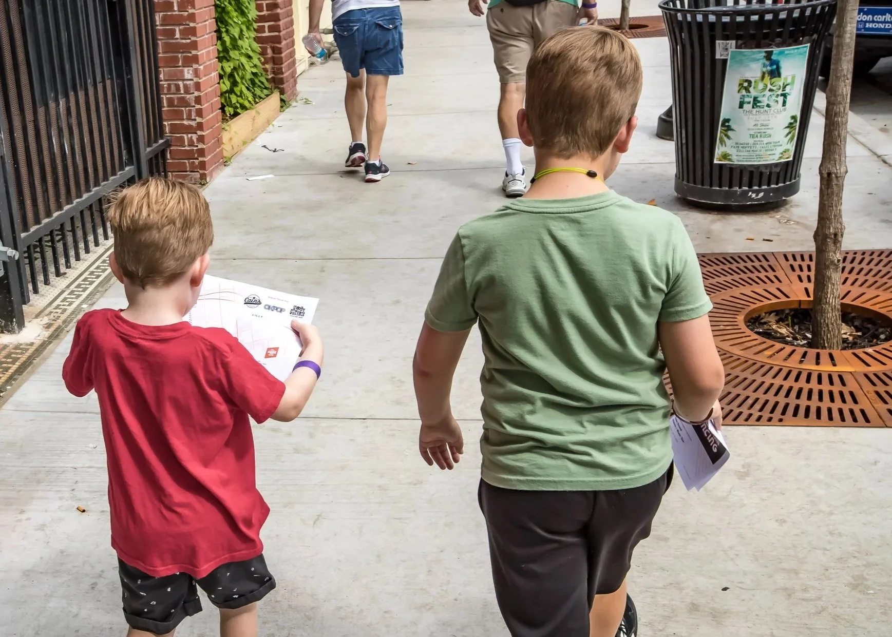 Two young boys walking side by side on a sidewalk, with one boy holding a map and the other holding the edge of a paper, near a tree and a trash can with a sign that reads 'Rush Fest.' Other people walk ahead in the background.