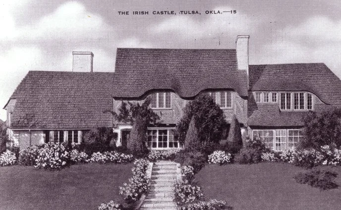 Black and white photograph of the Irish Castle in Tulsa, Oklahoma, showing a large house with a steep roof, multiple chimneys, surrounded by a well-maintained garden with flowers and a stone pathway leading to the entrance.