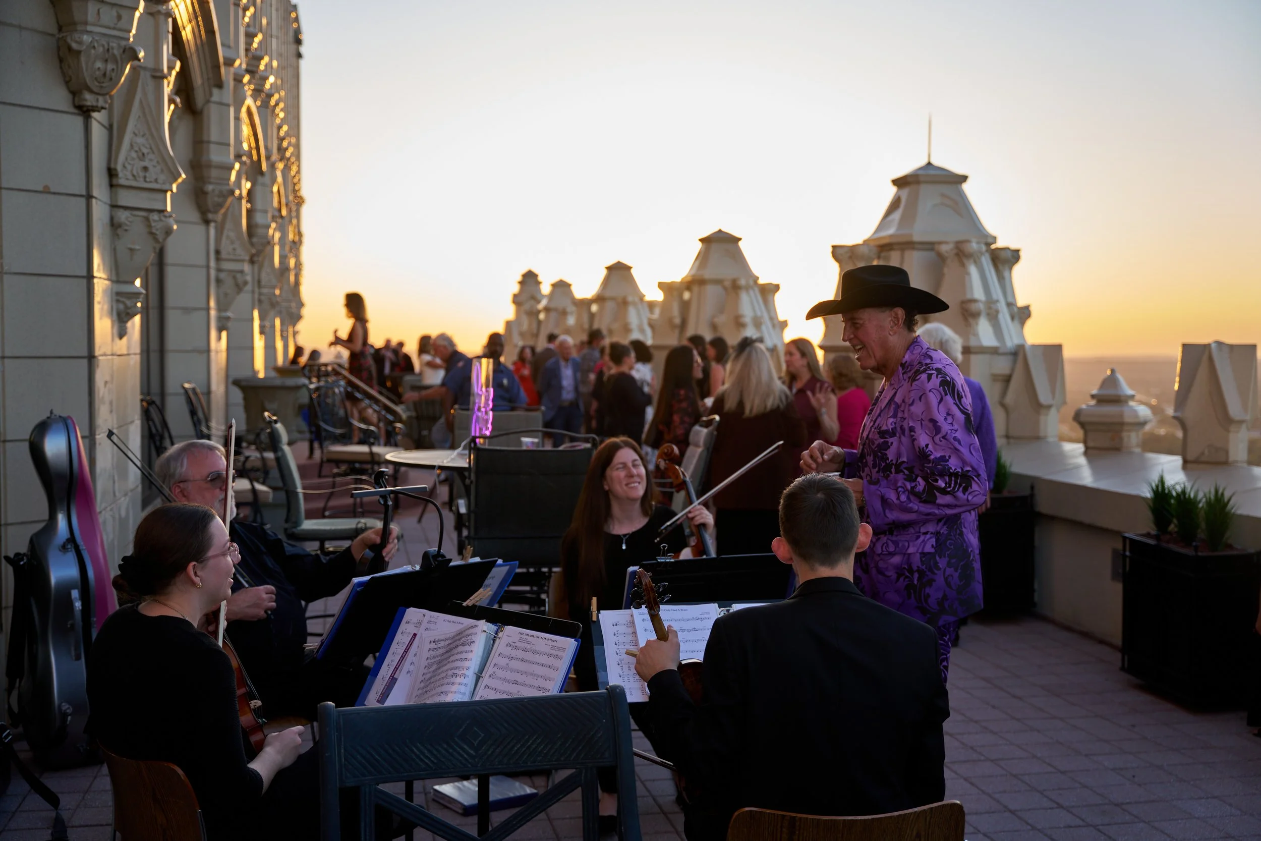 An outdoor orchestra performing at sunset on a rooftop terrace with architectural details and a city view in the background.