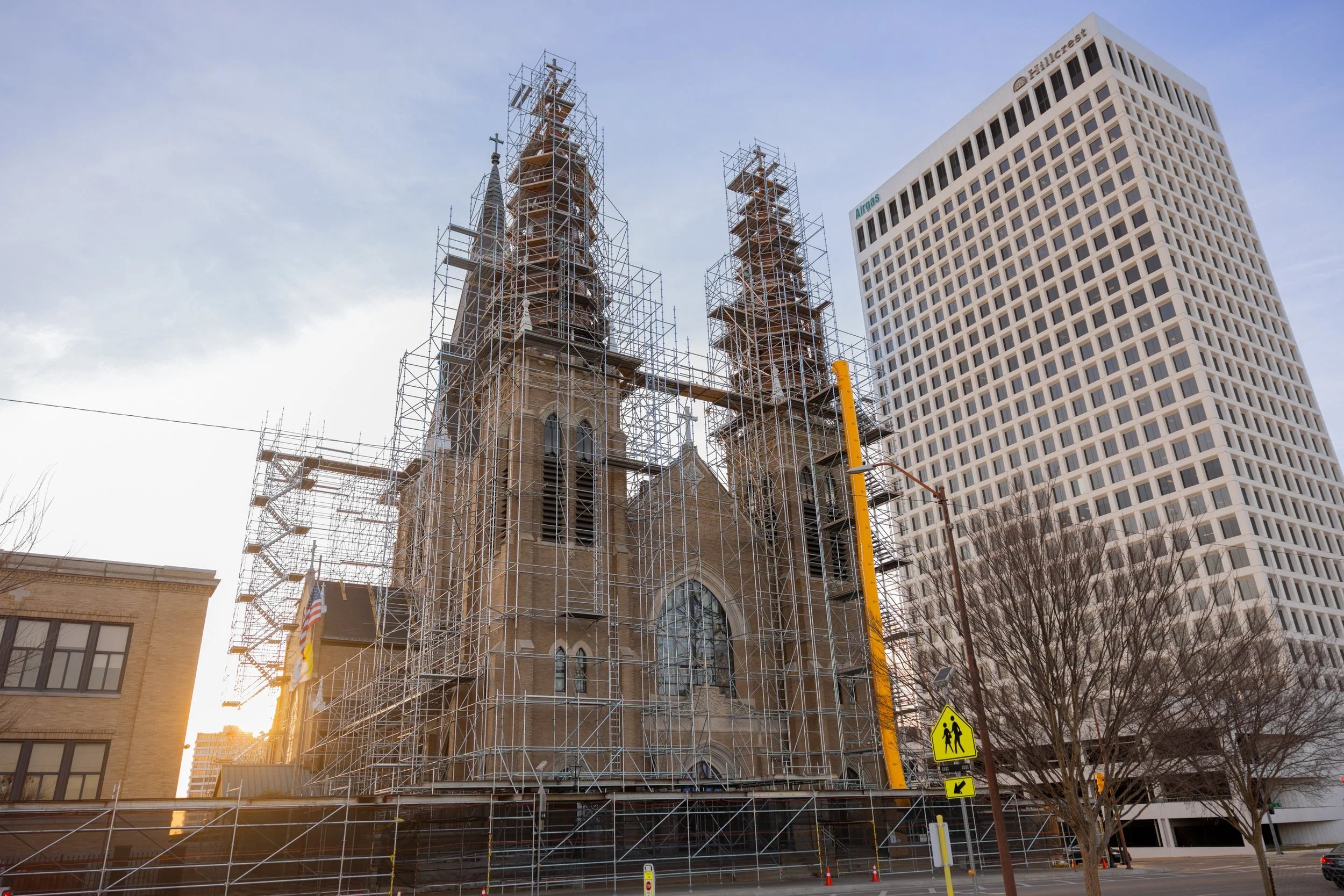 A historic church building with two tall steeples under renovation, surrounded by scaffolding, near modern high-rise buildings, at sunset.