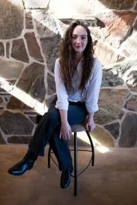 A young woman with long curly hair sitting on a black stool in front of a stone wall.