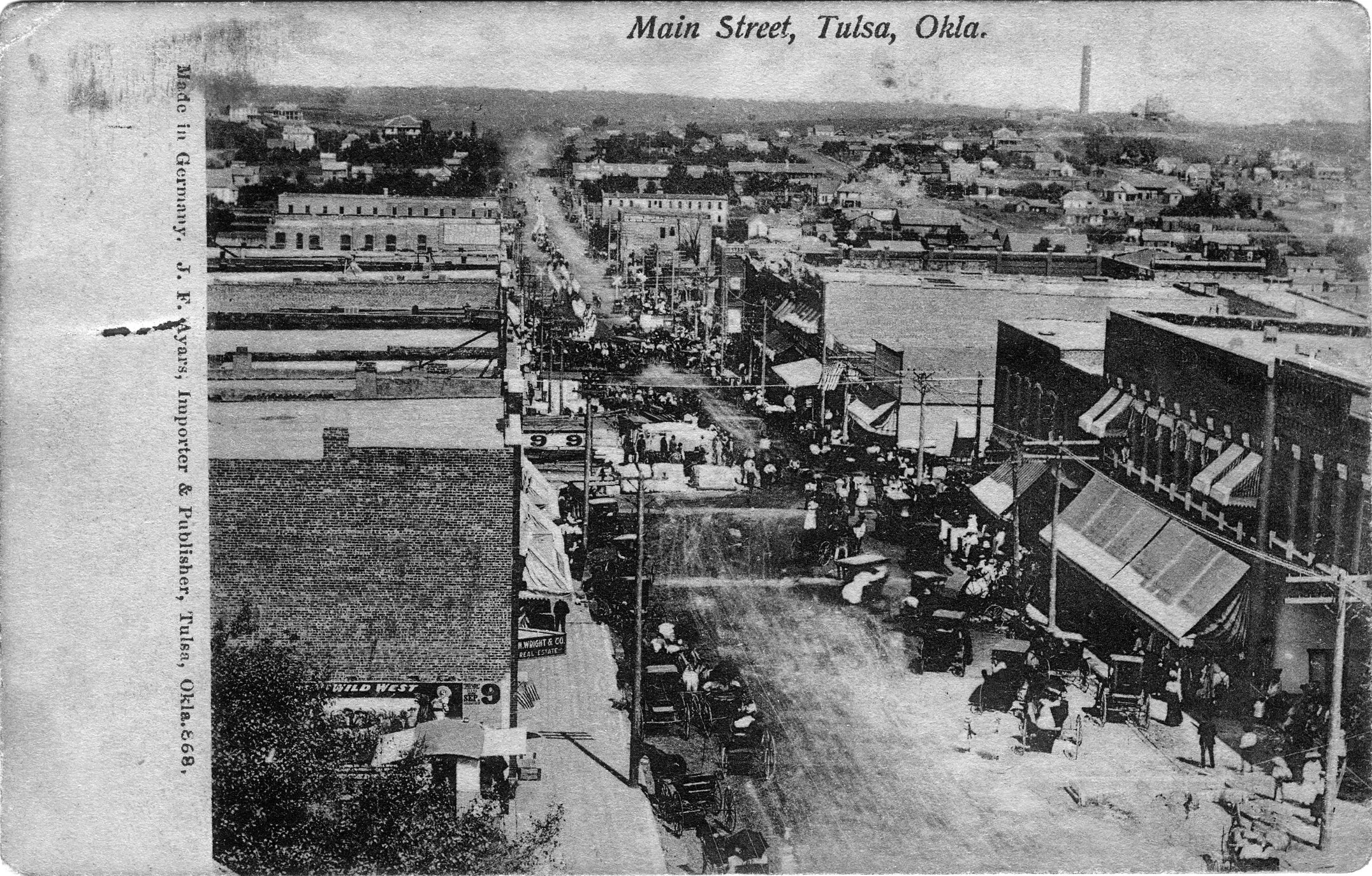 Historical black and white photograph of Main Street in Tulsa, Oklahoma, circa early 1900s, featuring vintage cars, horse-drawn carriages, and 19th-century storefronts with awnings.