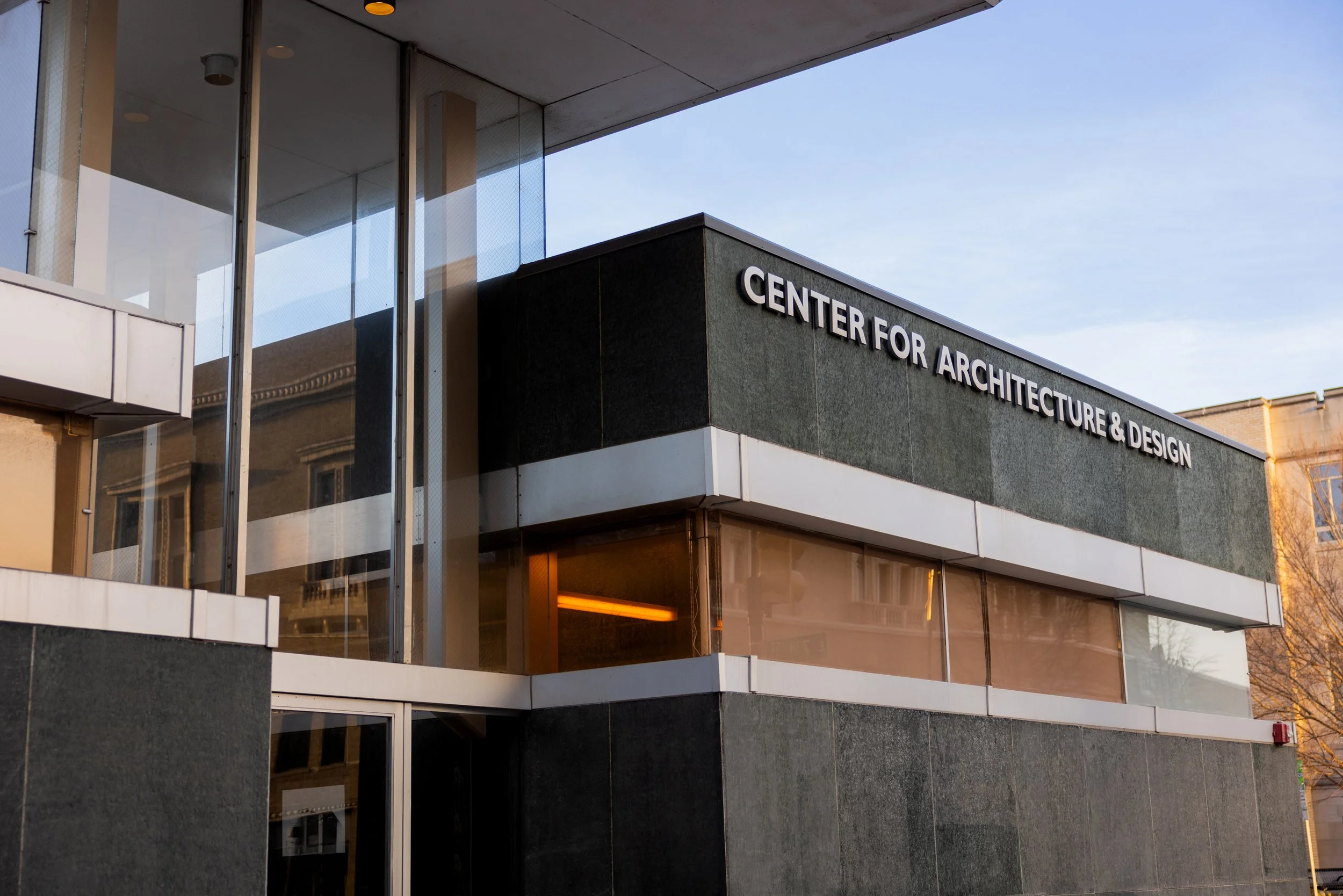 Exterior of a modern building with glass and dark stone facade, displaying a sign for the Center for Architecture & Design.