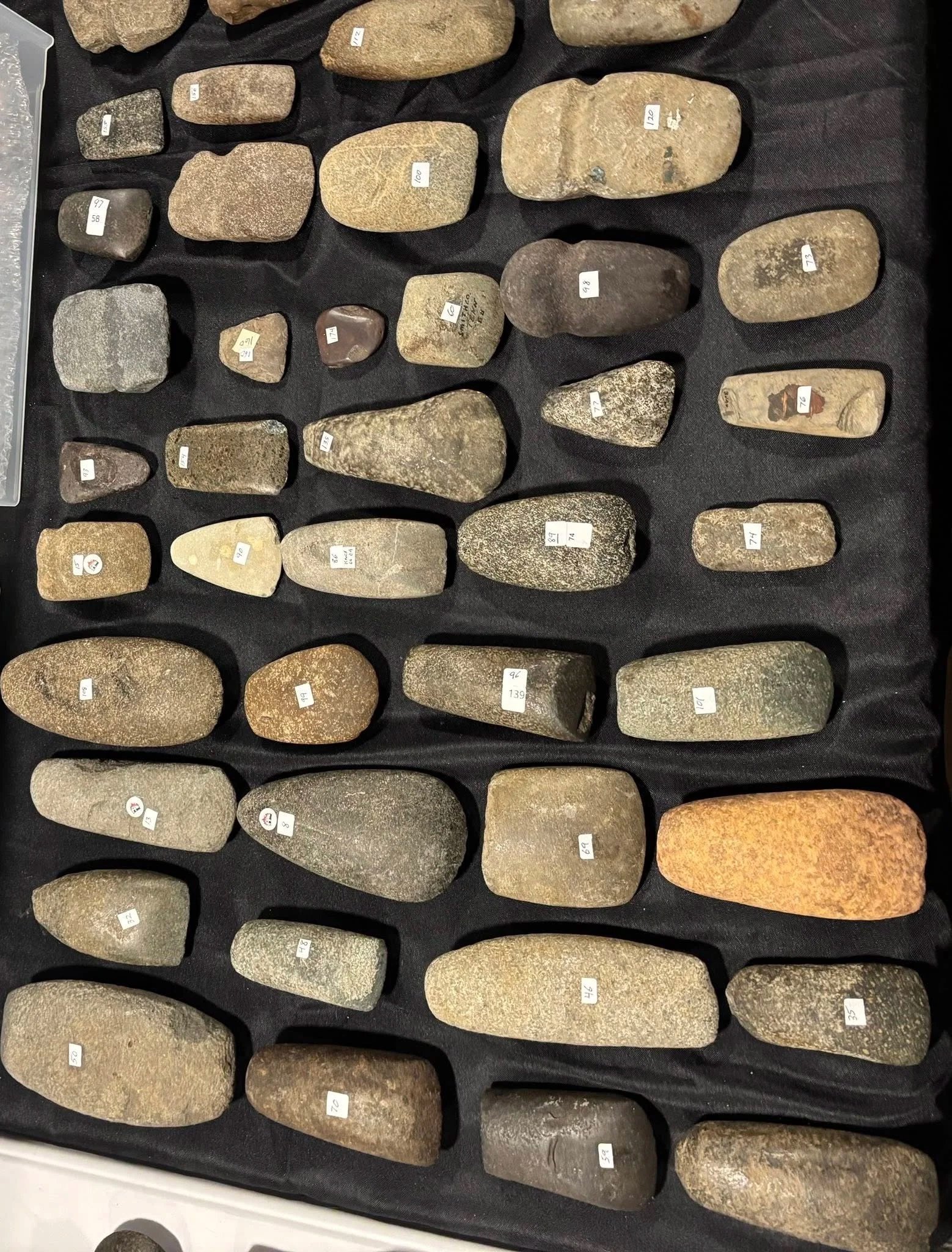 Collection of various ancient stone tools and artifacts displayed on a black cloth, each labeled with a small white sticker.
