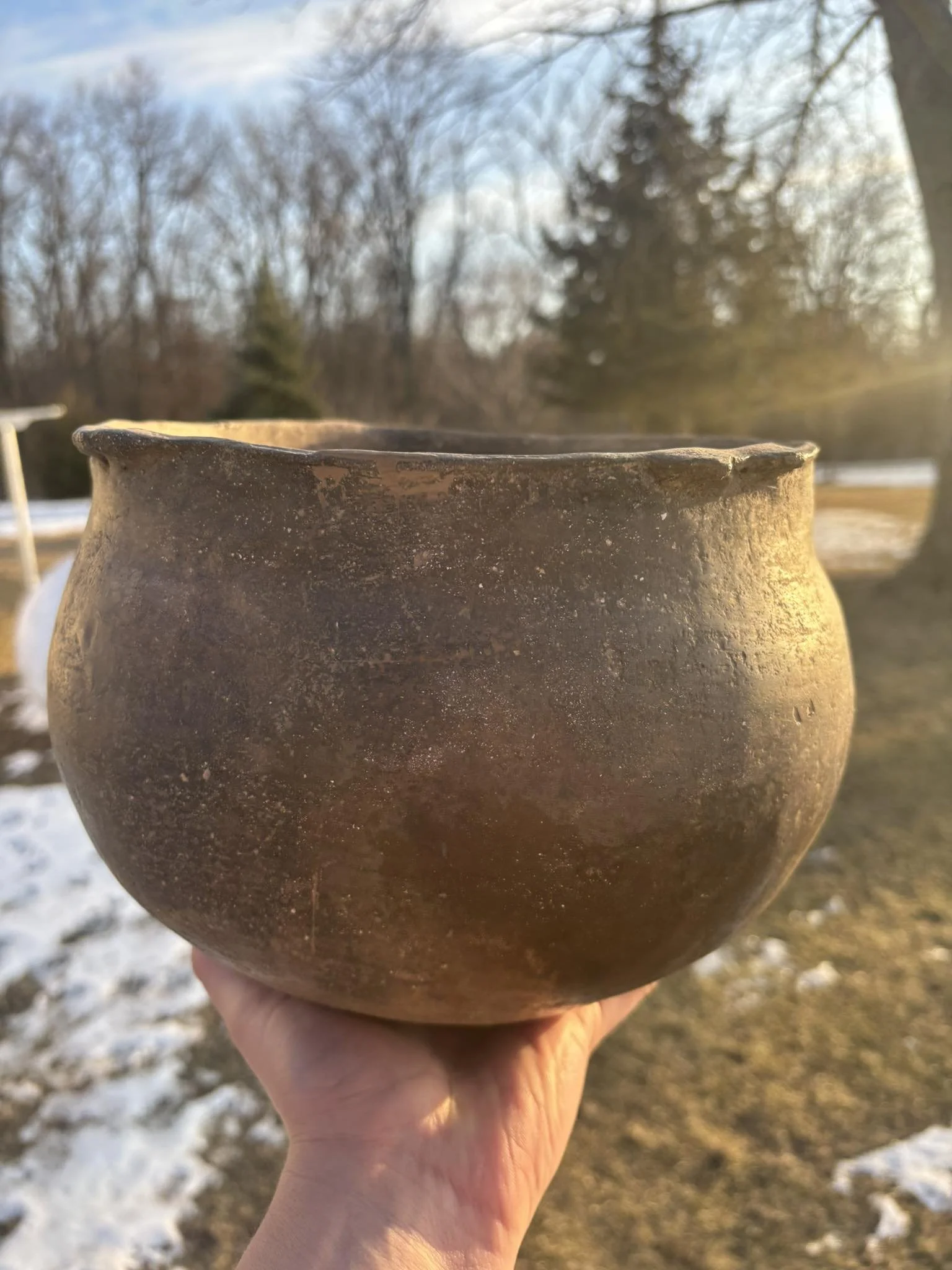 Close-up of an aged clay pot held outside during sunset, with trees and a partly cloudy sky in the background.