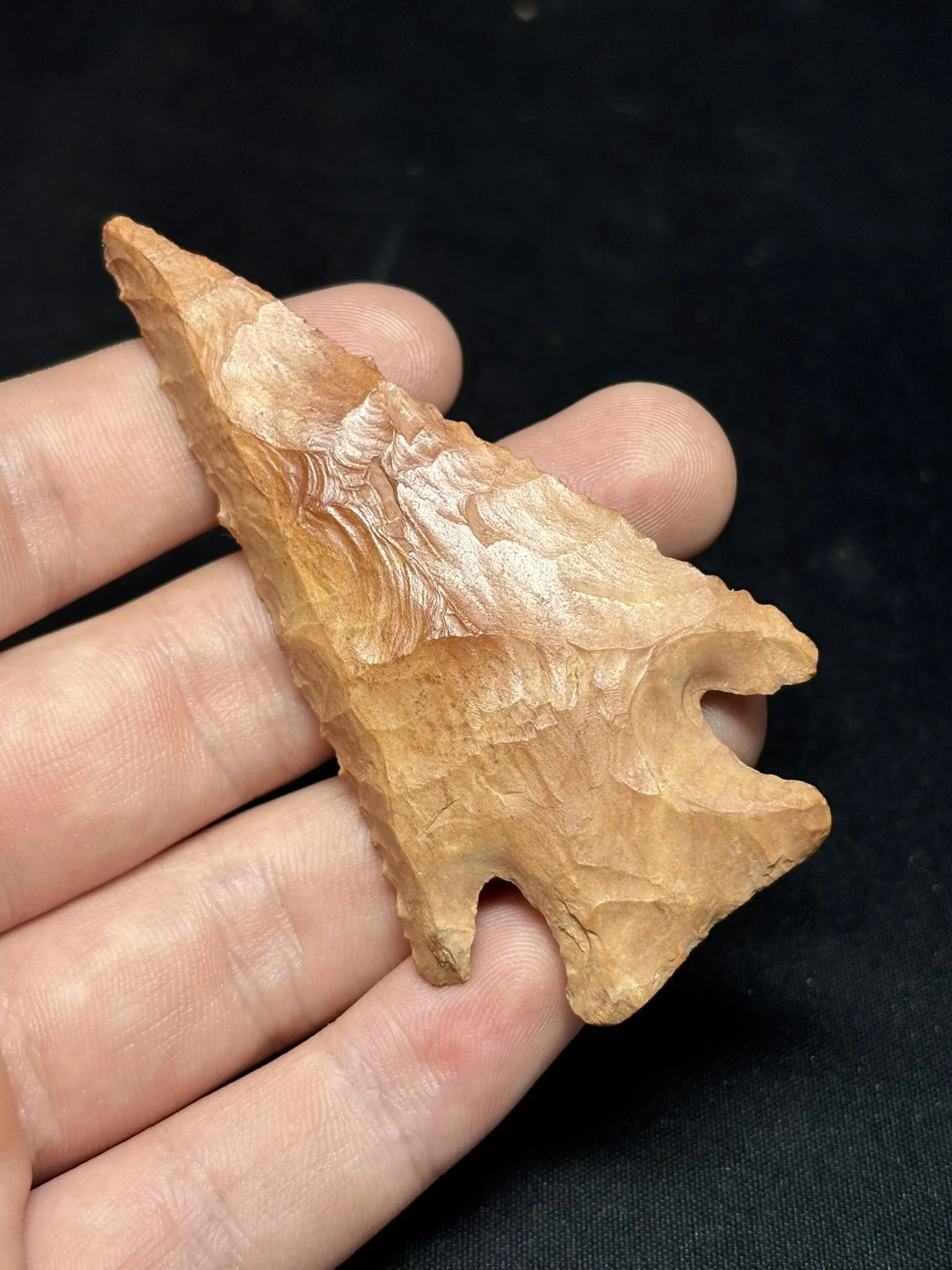 Close-up of a person's hand holding a large, flaked, orange-brown stone arrowhead against a black background.