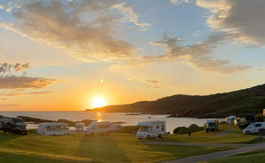 Sunset over a coastal campground with several camper vans parked on green grass and a scenic view of the ocean and hills in the background.