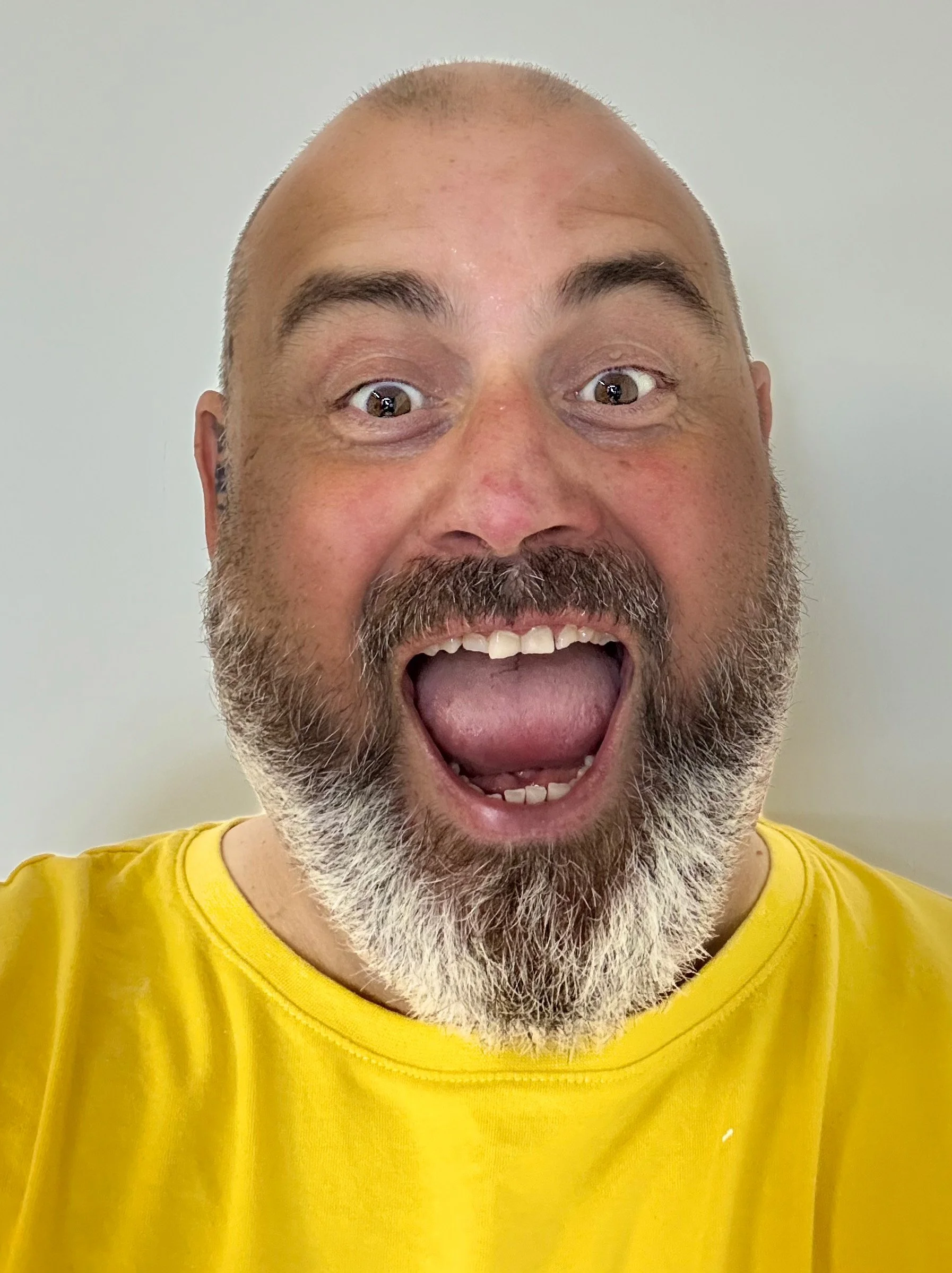 A man with a beard and mustache, bald head, wearing a yellow shirt, smiling widely with his mouth open, against a plain background.